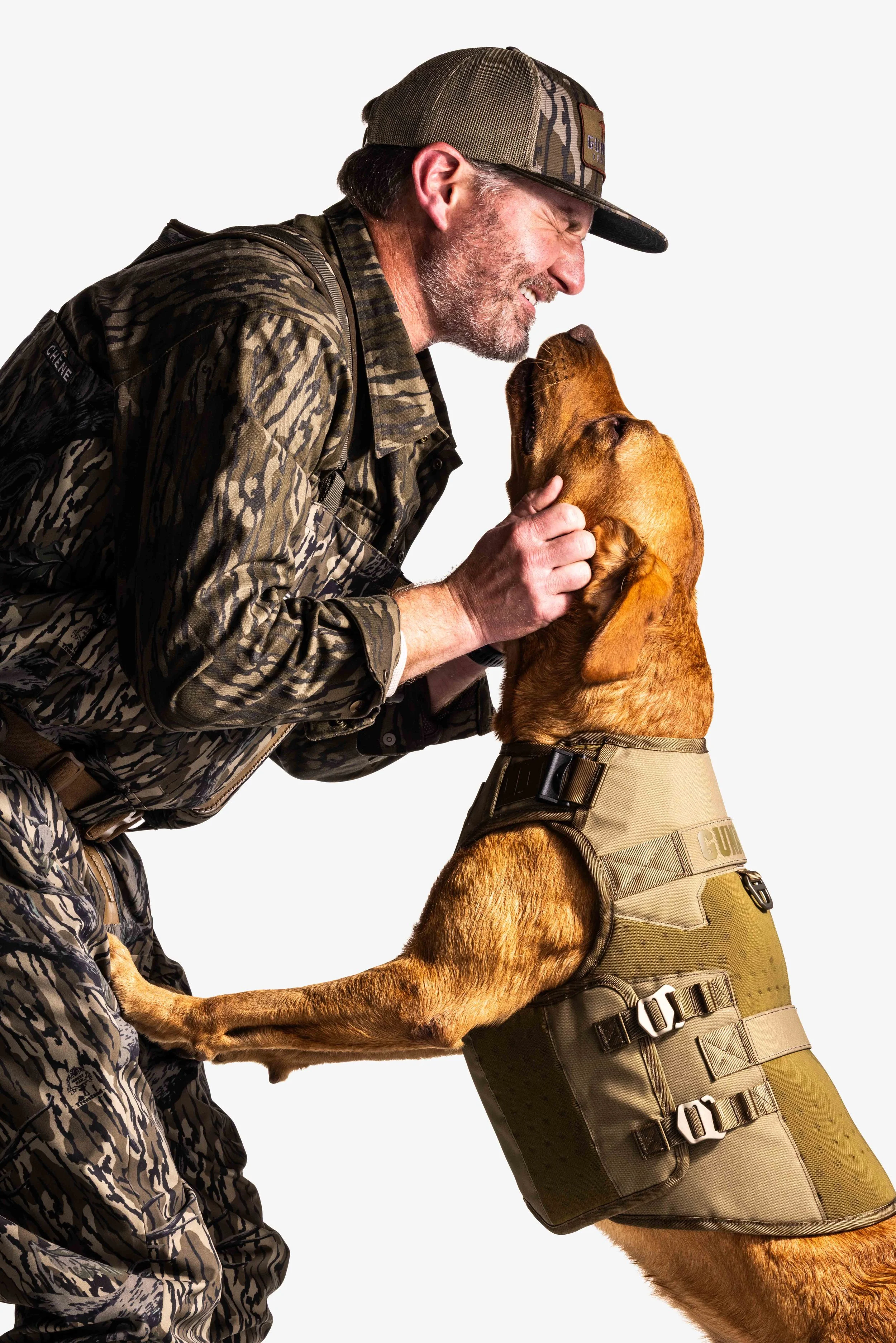 A man and a dog are facing each other, with the man smiling and the dog looking up at him. The man is wearing camouflage clothing and a cap, and the dog is wearing a protective vest.