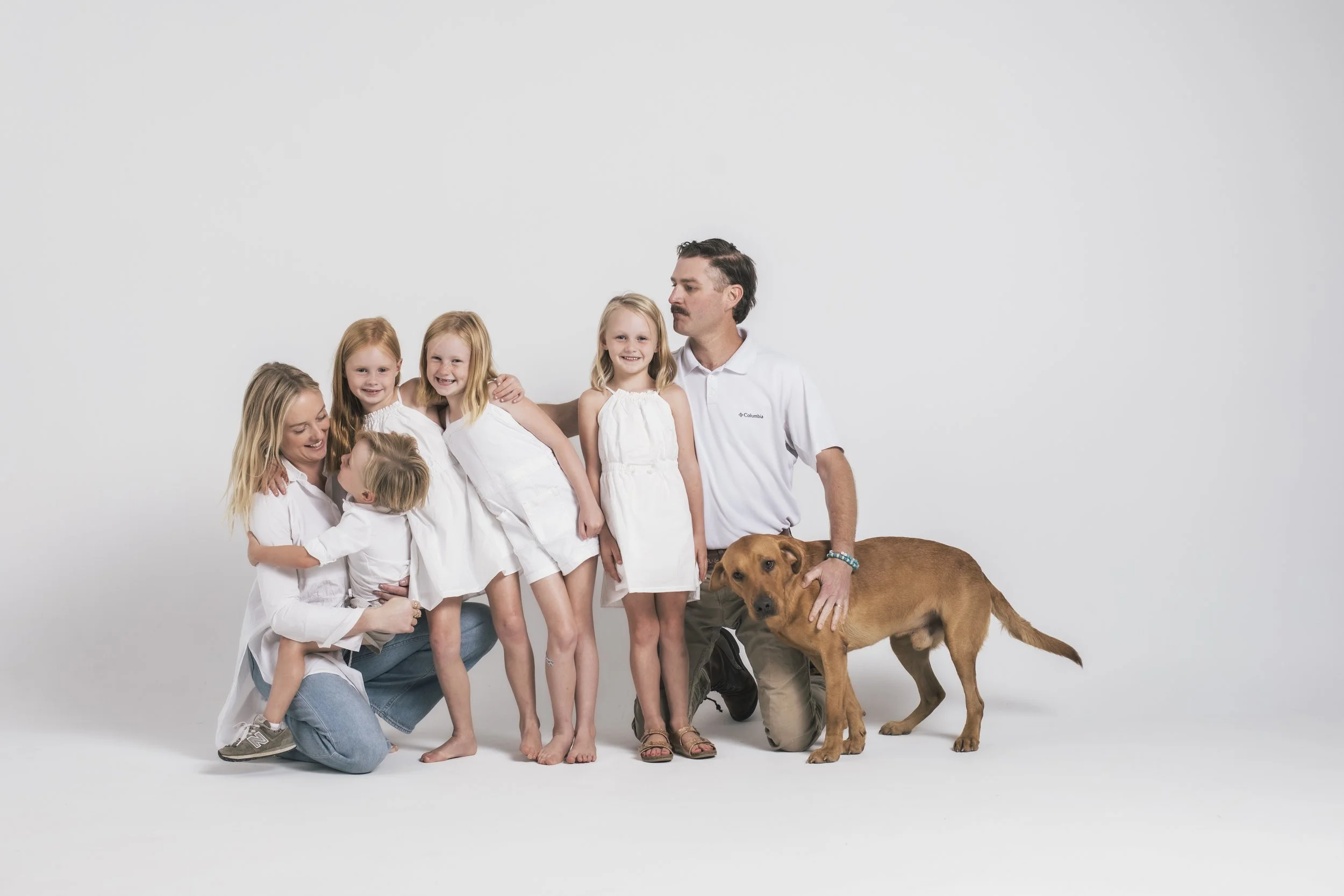 A family of seven people, including five young girls, an adult woman, an adult man, and a large brown dog, posing together in a studio with a plain white background.