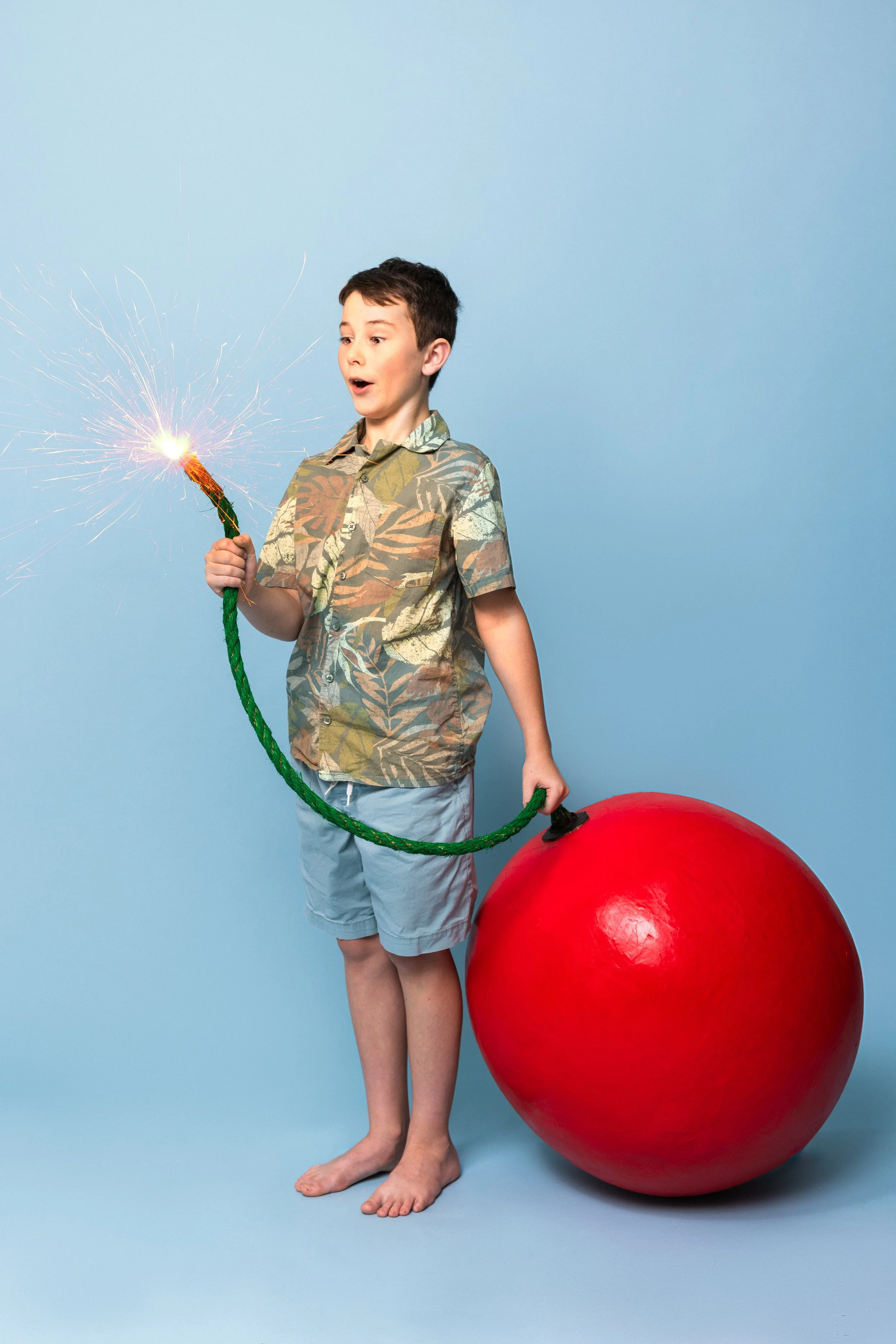 A young boy in a Hawaiian shirt and shorts holding a sparkler in one hand and a large red ball with a fuse in the other, standing barefoot on a light blue background.