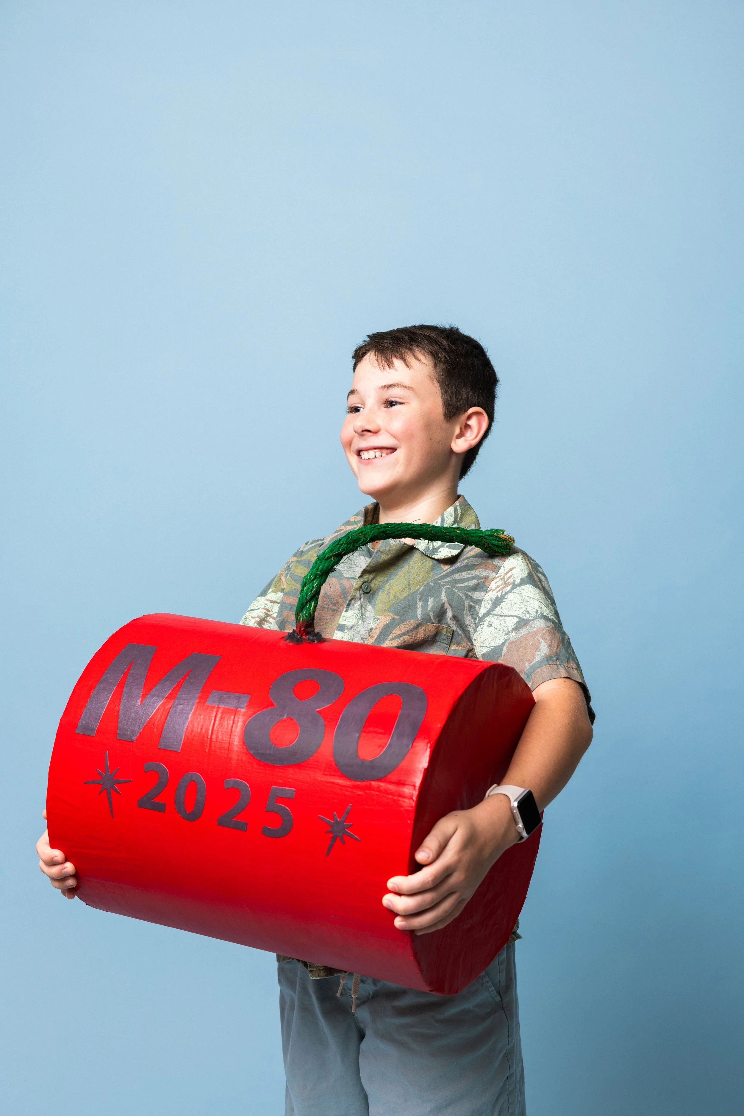 A smiling boy holding a red model of a missile with M-80 and 2025 written on it in black letters, standing against a plain light blue background.
