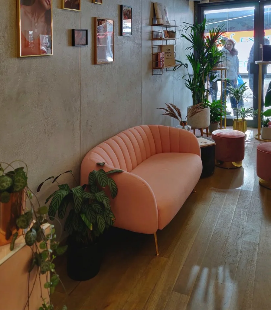 Cozy cafe interior with a pink velvet sofa, multiple potted plants, small side tables, and artwork on the wall. A woman is visible outside through the glass door.