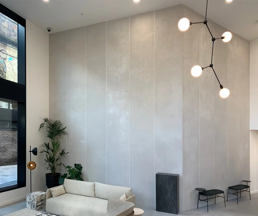 Modern living room with large window, beige sofa, potted plants, marble side table, black wall-mounted lamp, contemporary black stool, and pendant light fixture with spherical bulbs.