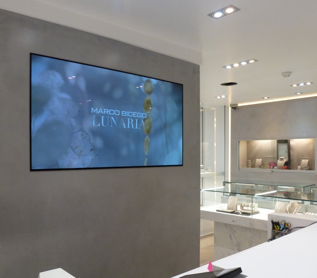 A jewelry store interior with a large screen displaying 'Marco Bicego Lunaria' against a gray wall, jewelry displays with necklaces and accessories in glass cases, and bright ceiling lights.