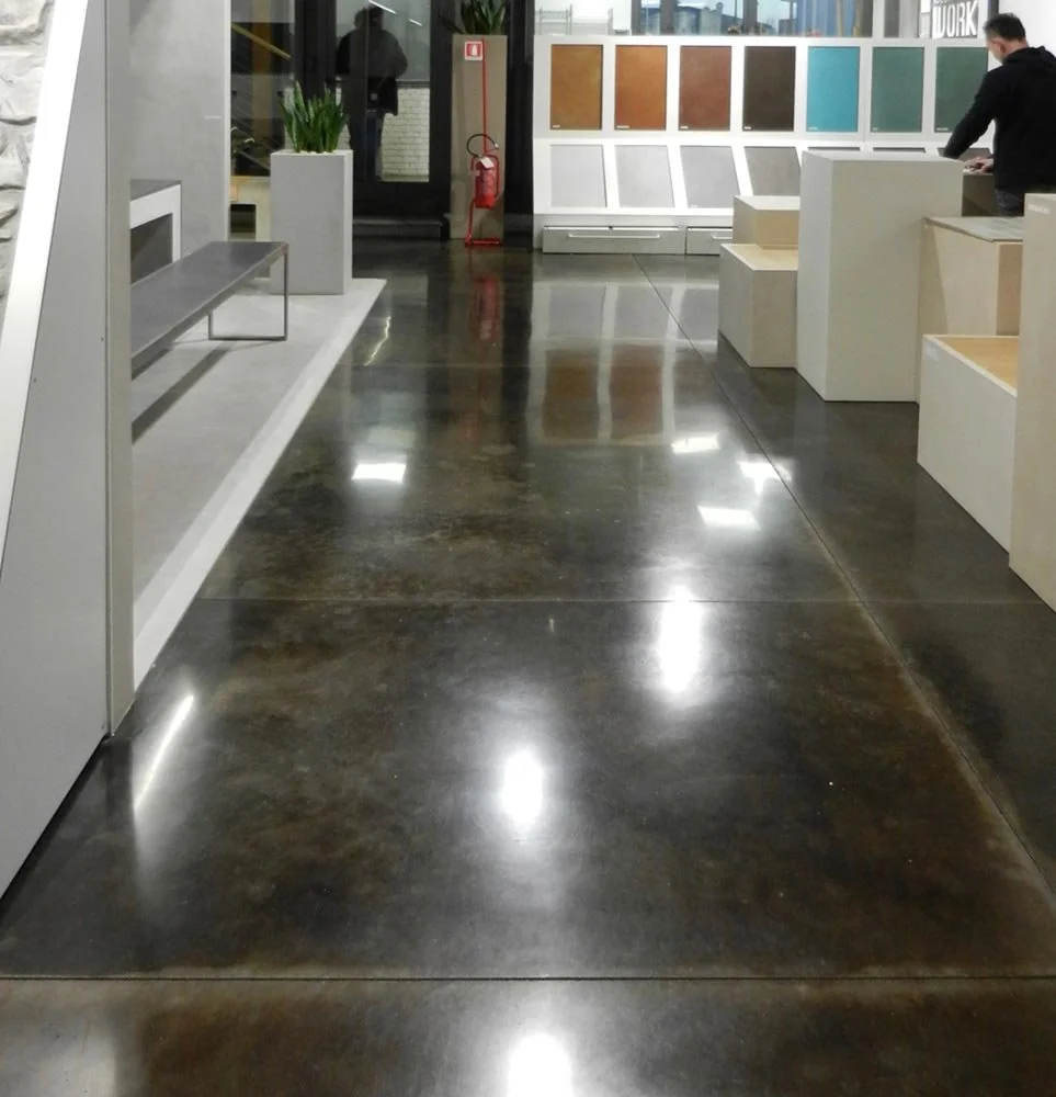 Interior of a modern showroom or store with a glossy polished concrete floor, display samples of paint colors on the wall, a bench, potted plant, and a person browsing.