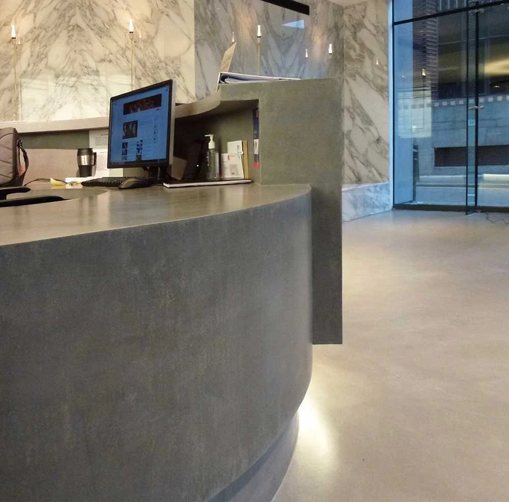 Office reception desk with a computer monitor, a travel mug, and some documents, with marble walls and glass doors in the background.