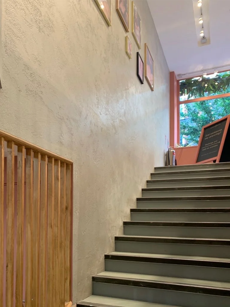 Indoor staircase with wooden handrail and framed art on beige textured wall leading toward a window with greenery outside.