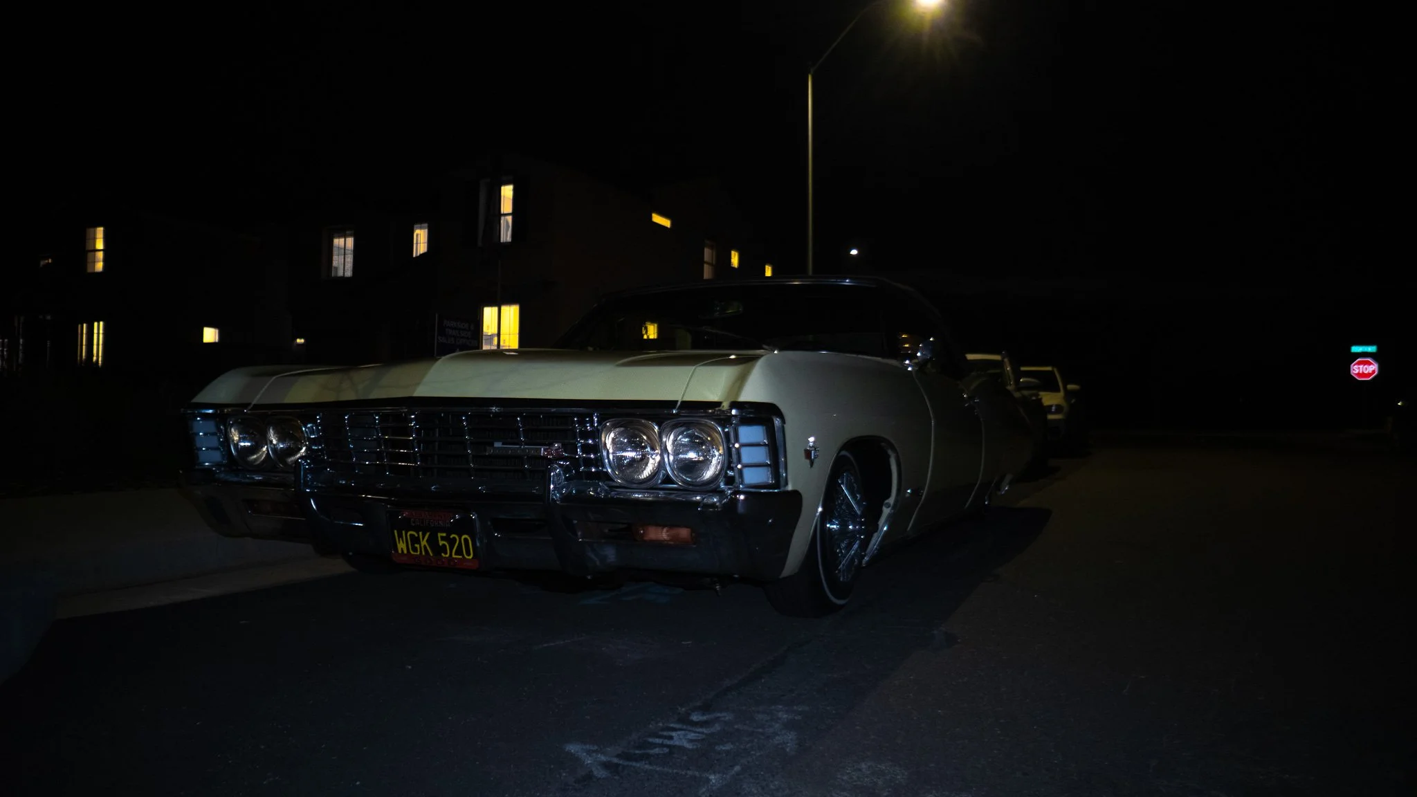 A vintage white car parked on a dark street at night with a building in the background, illuminated windows, and a streetlight overhead.