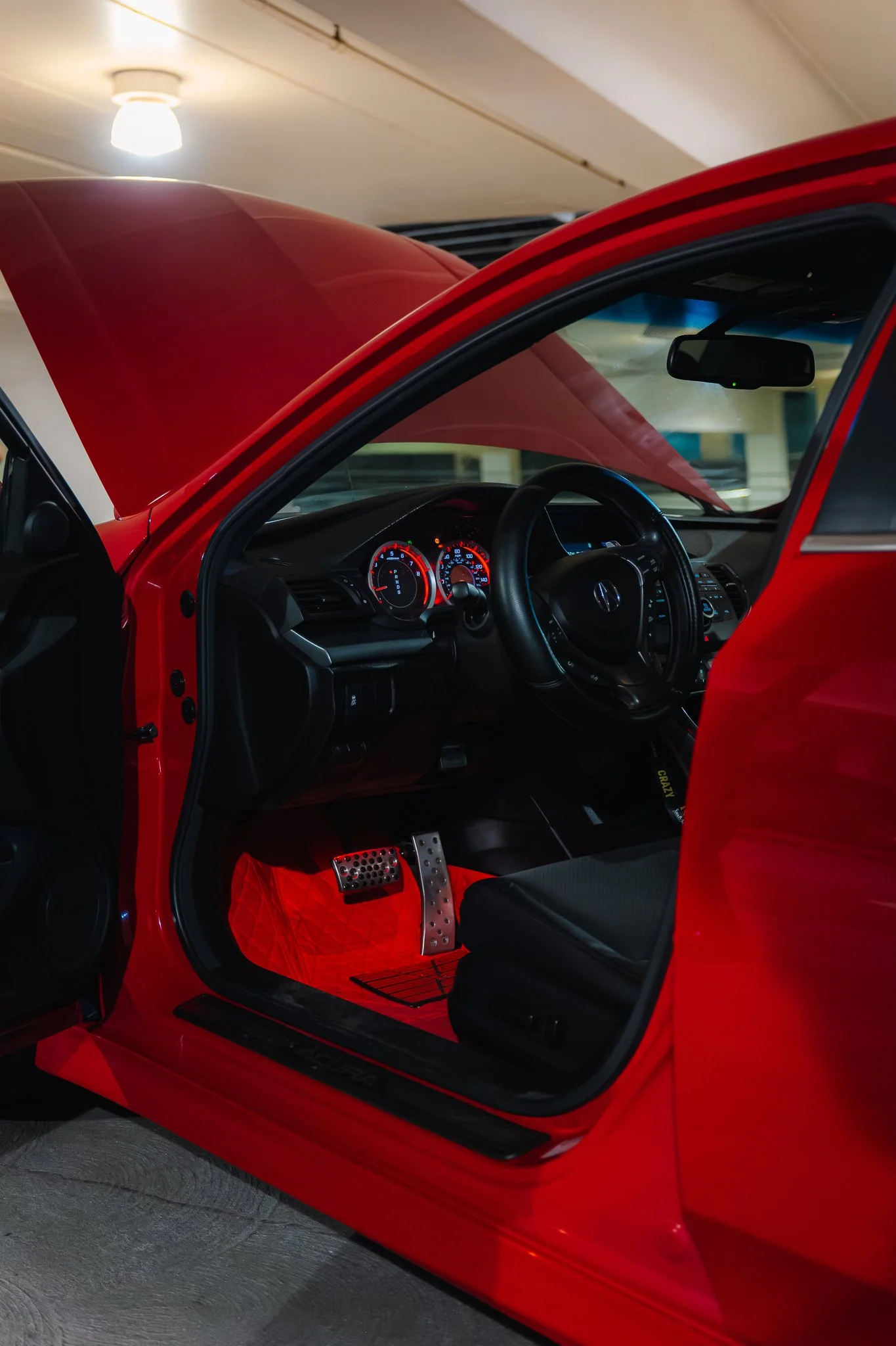 Close-up of a red sports car interior with a black steering wheel, dashboard with illuminated gauges, and red floor mats, seen from the passenger side with the door open.