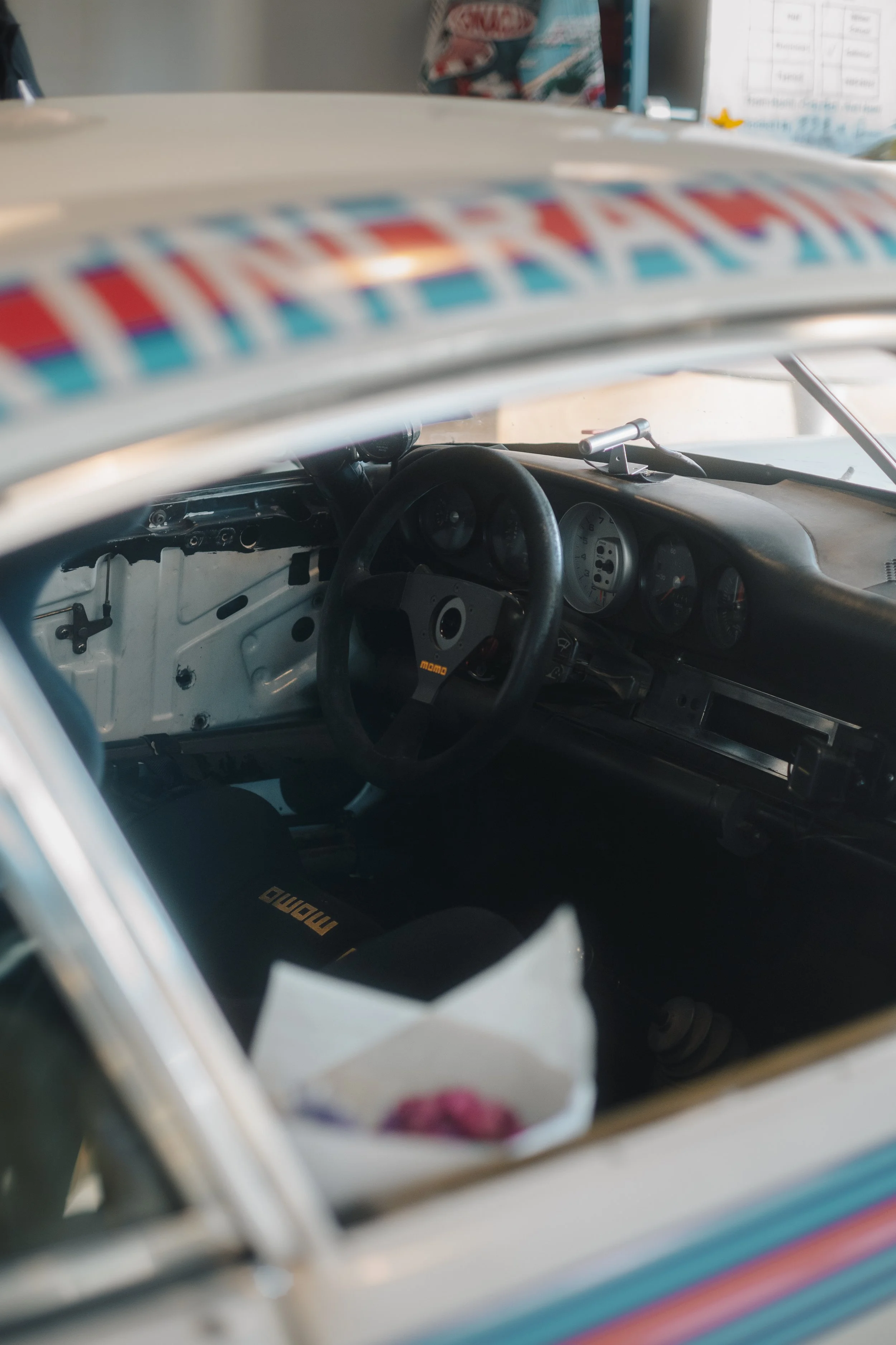 View through the window of a vehicle showing the dashboard and interior, with the dashboard featuring several gauges and a black steering wheel, and the interior including a racing seat with the brand name 'MOMO' visible.