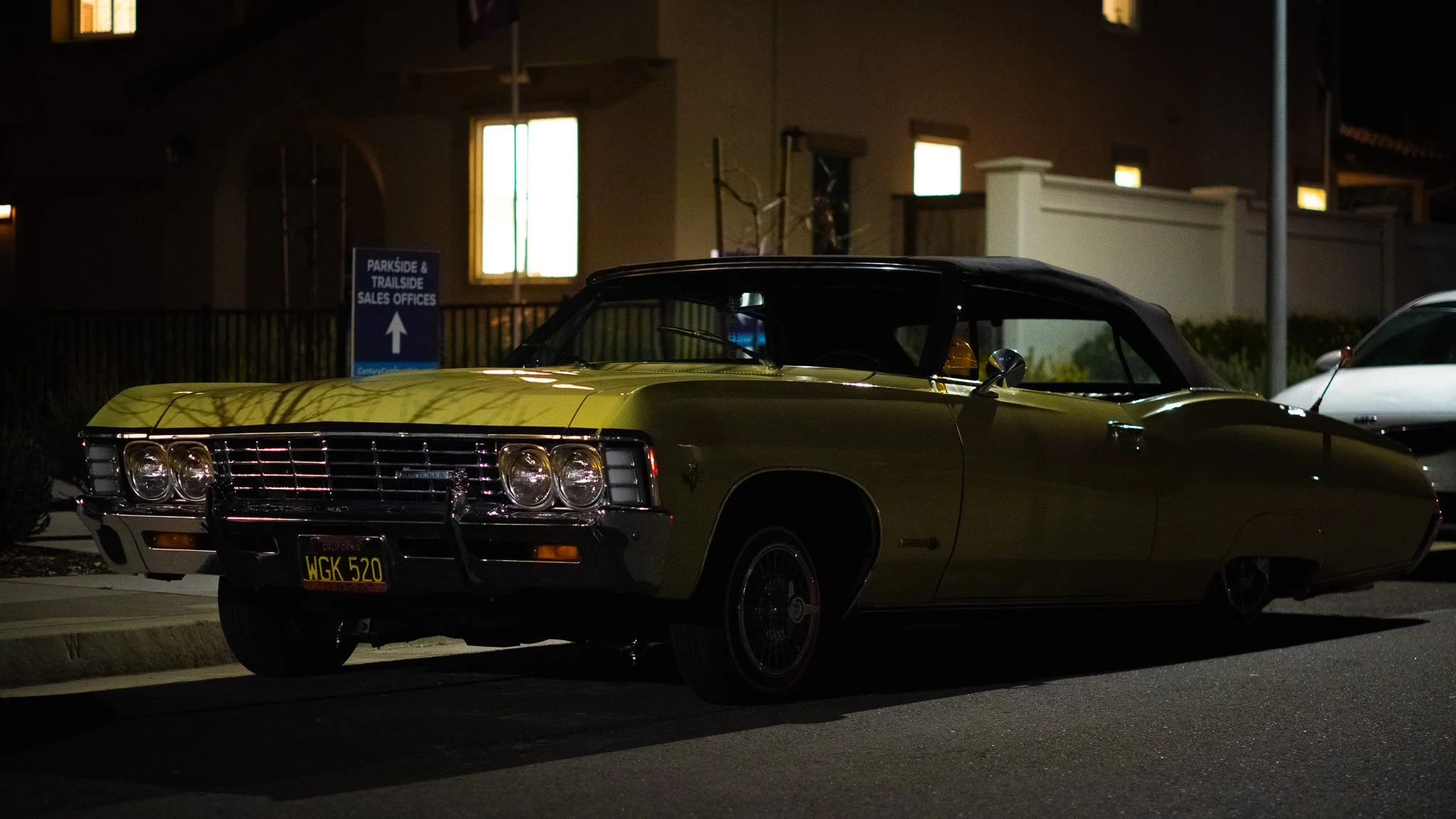 A vintage yellow convertible car parked on the street at night with a building in the background and a sign indicating sales offices for parkside and trailside.