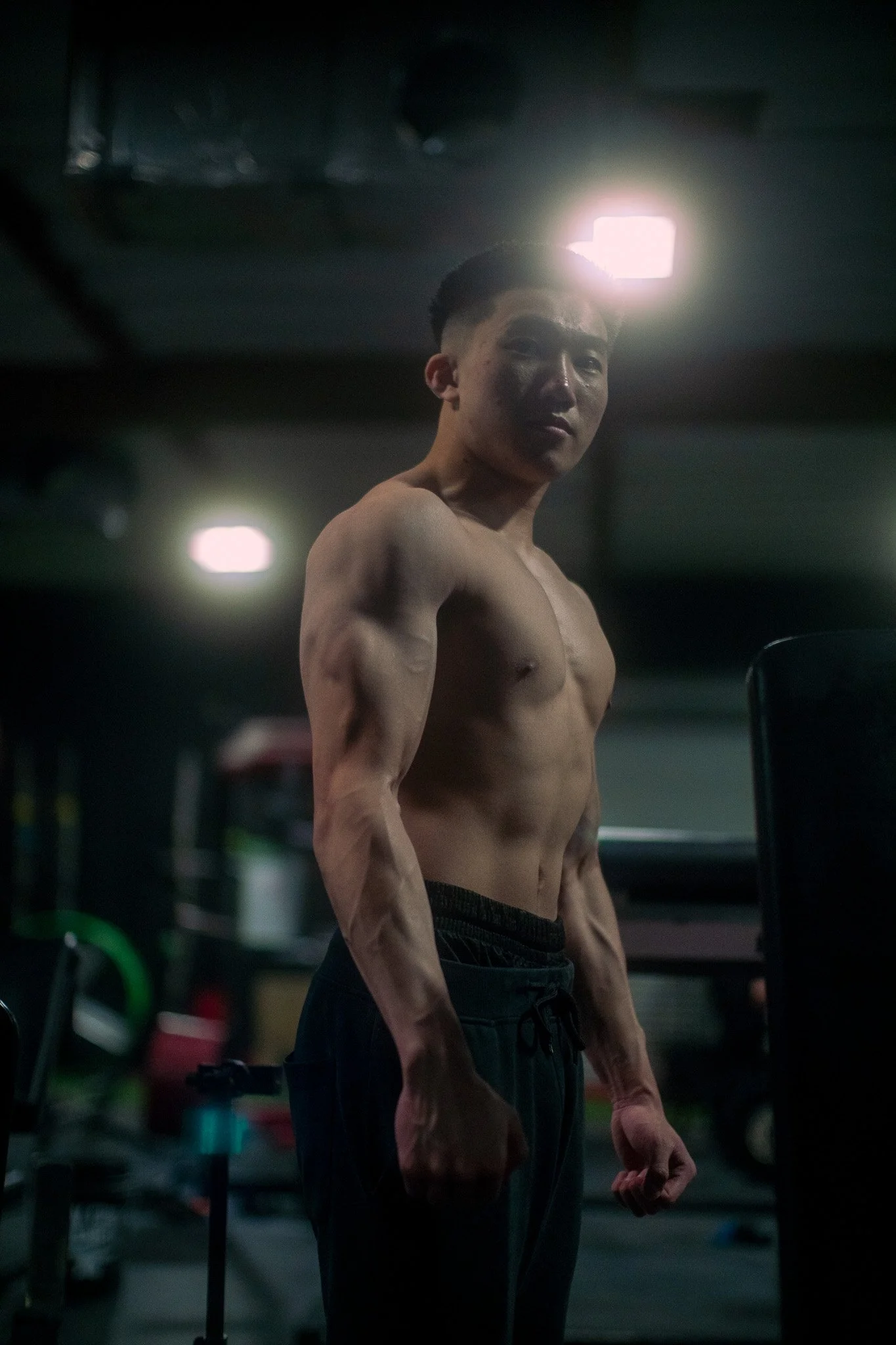 A shirtless young man with muscular build and dark hair standing in a dimly lit gym, with a bright light source behind his head, and workout equipment visible in the background.