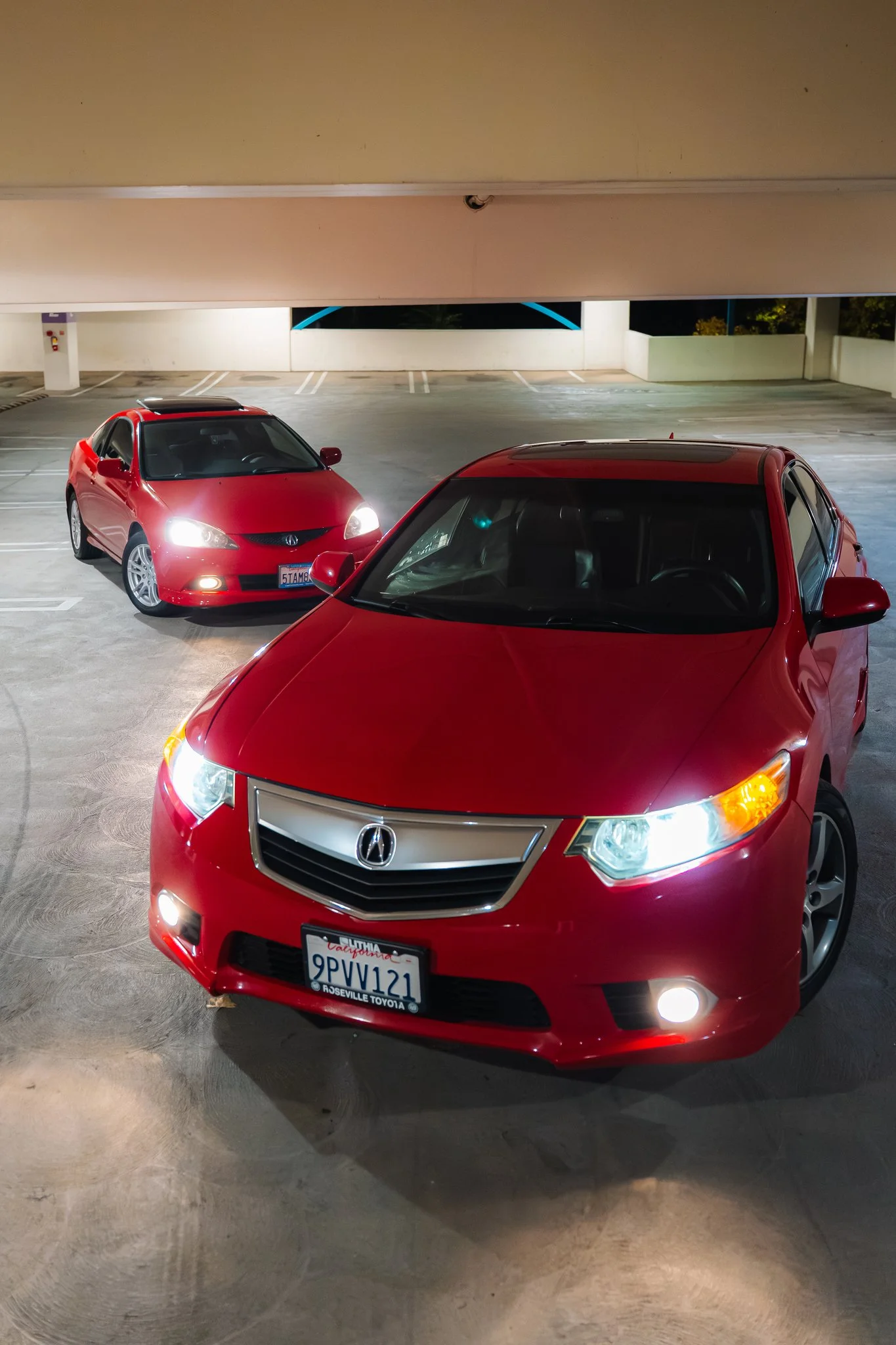 Red Acura sedan parked in an indoor parking garage at night, with the front of the car facing the camera. The car has a California license plate and is illuminated by overhead lights.