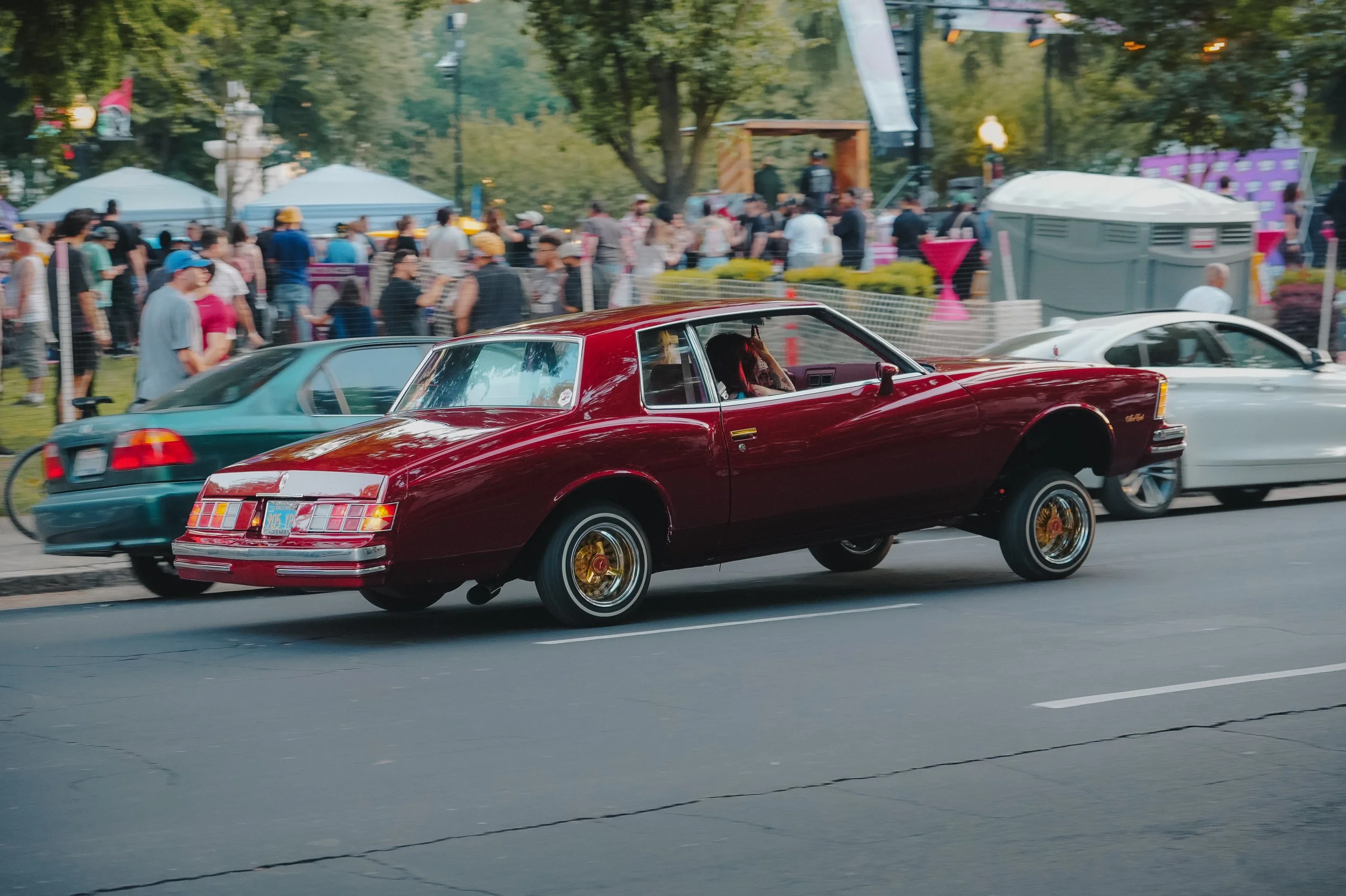 A red vintage car driving on a city street with a crowd of people in the background, possibly during a parade or event.