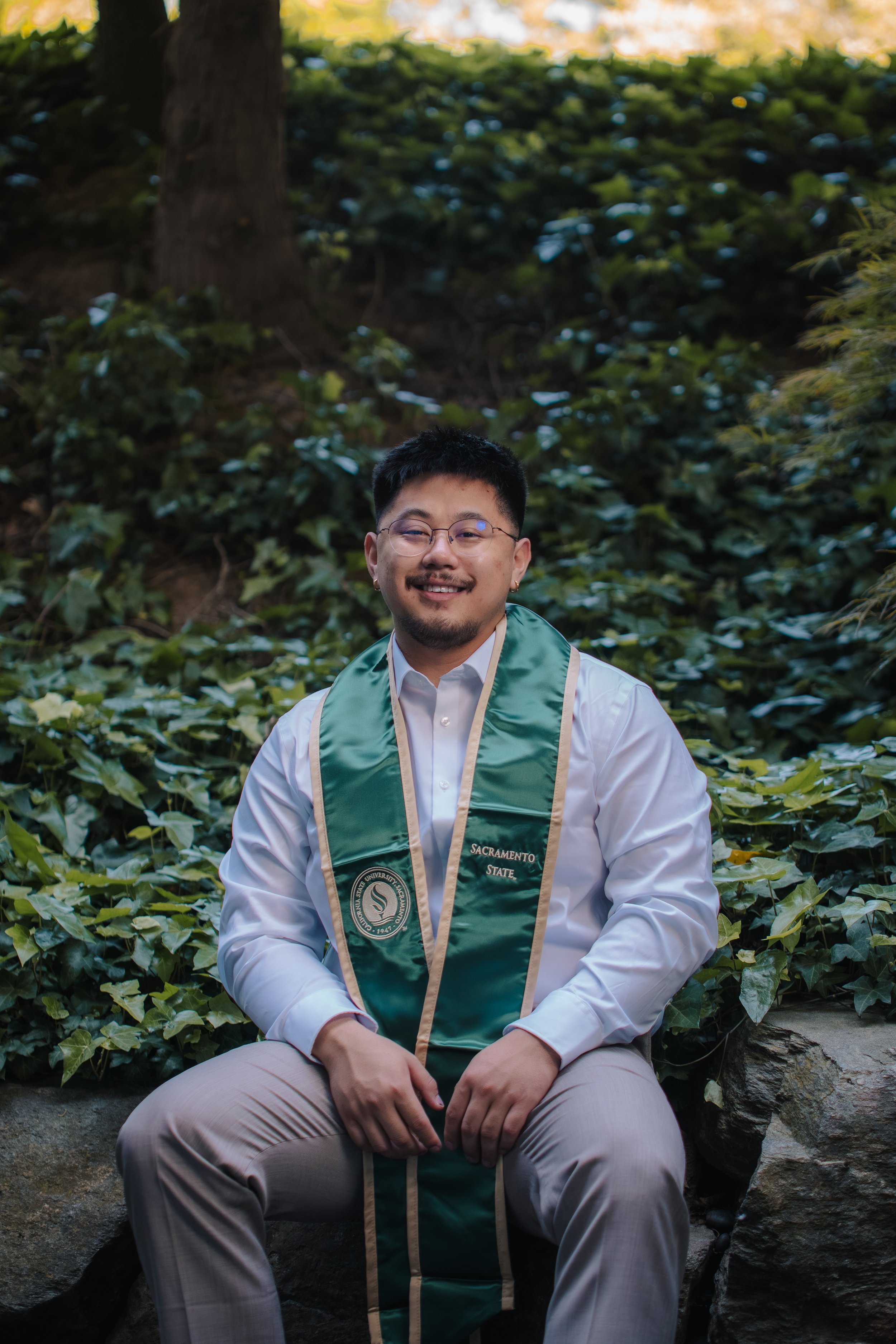 A man smiling, wearing glasses and a green Sacramento State graduation stole, sitting outdoors on a rock in front of a lush, green leafy background.