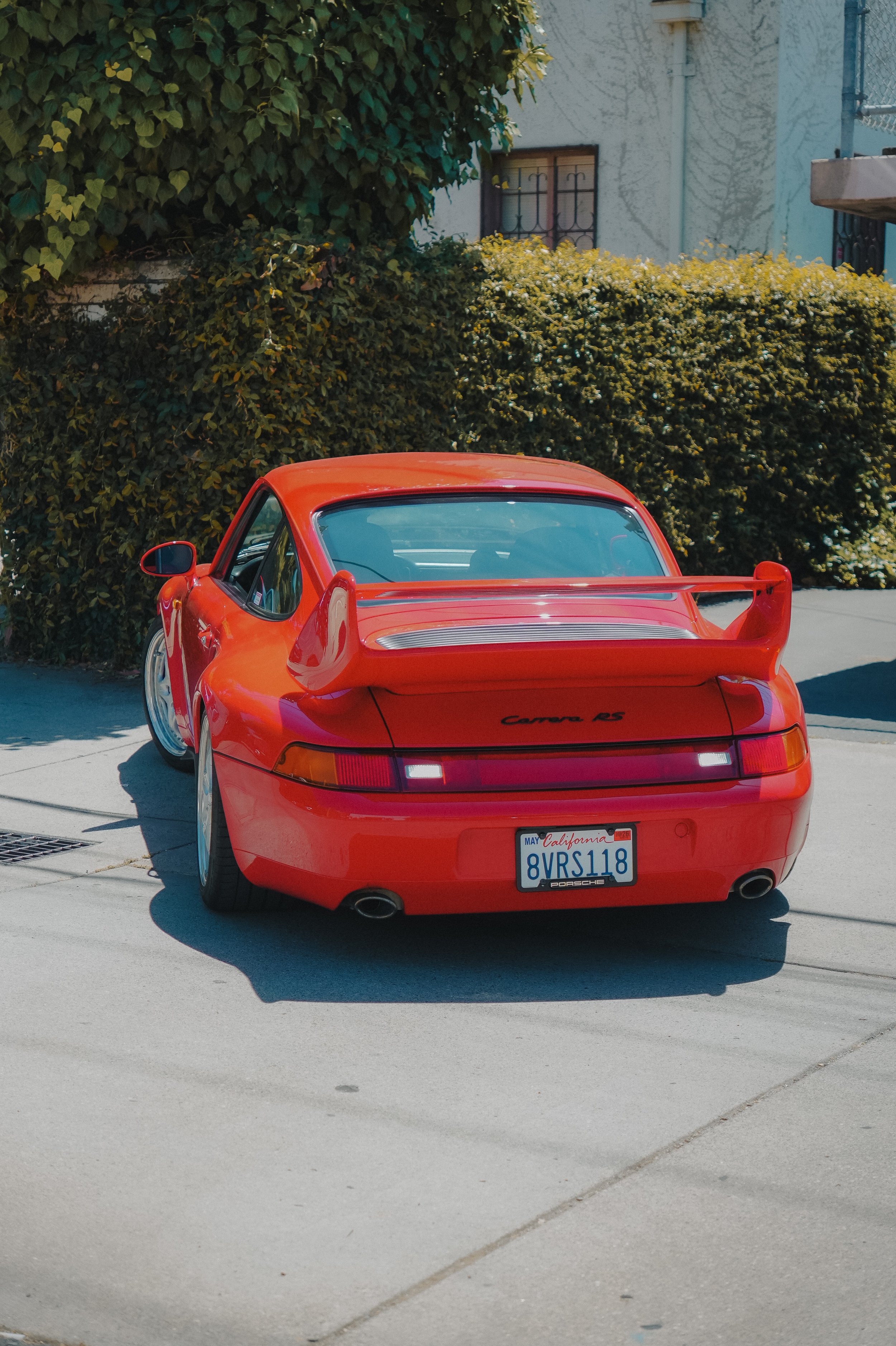 A red Porsche Carrera RS sports car parked on a driveway in front of a green hedge and a residential building with a window. The car has a large rear wing and a California license plate reading 8VRS118.