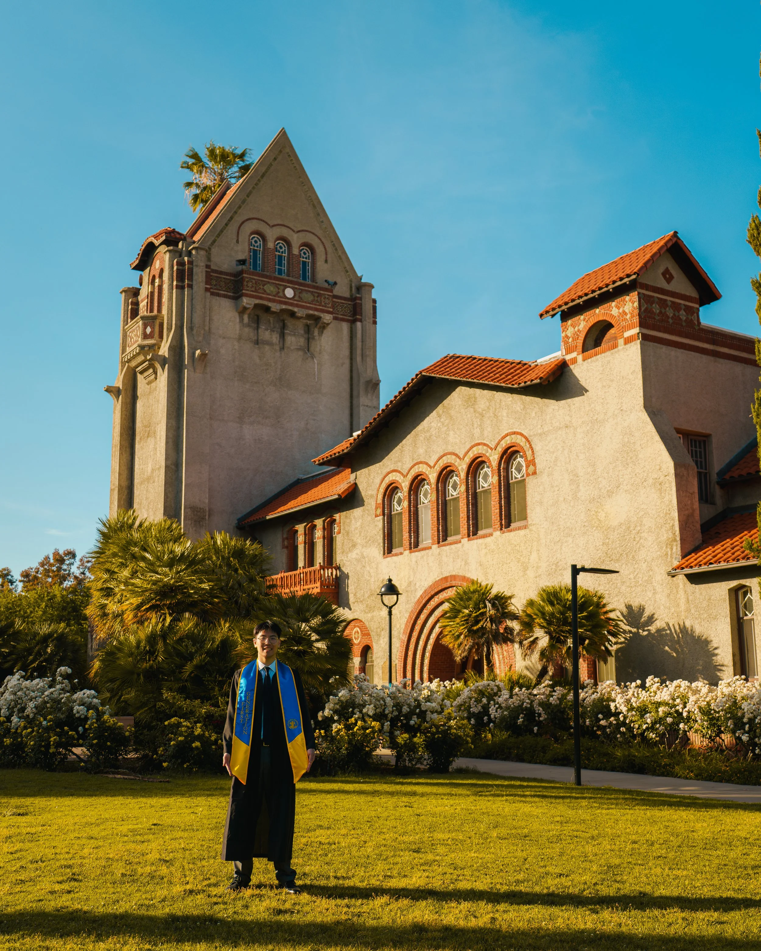 A person in a graduation gown and cap standing on a grassy area in front of a historic building with a tower and red tile roof, during golden hour with warm sunlight and a blue sky.