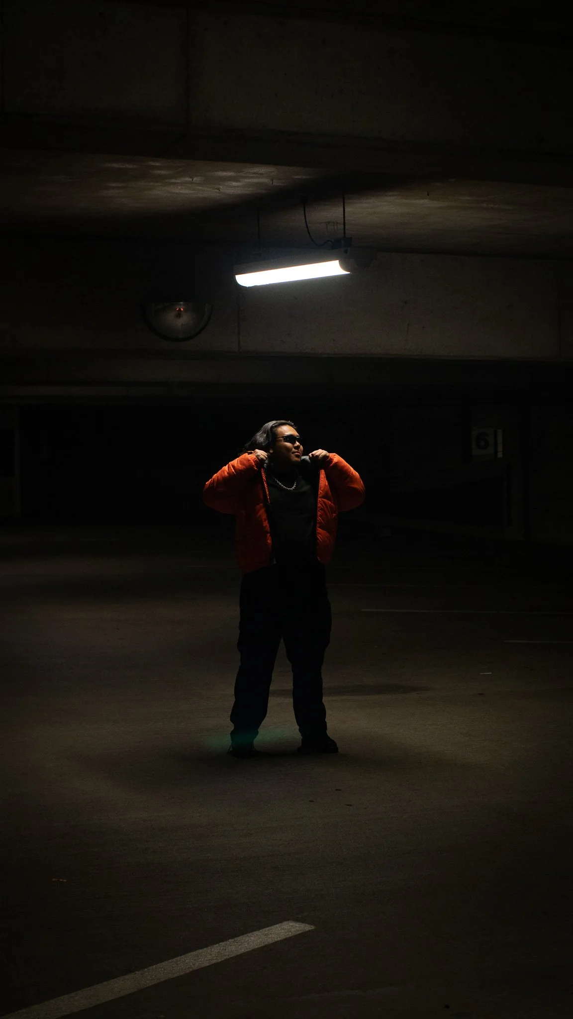 A person standing in a dark parking garage, wearing a red jacket, sunglasses, and black pants, with their hands on their shoulders.
