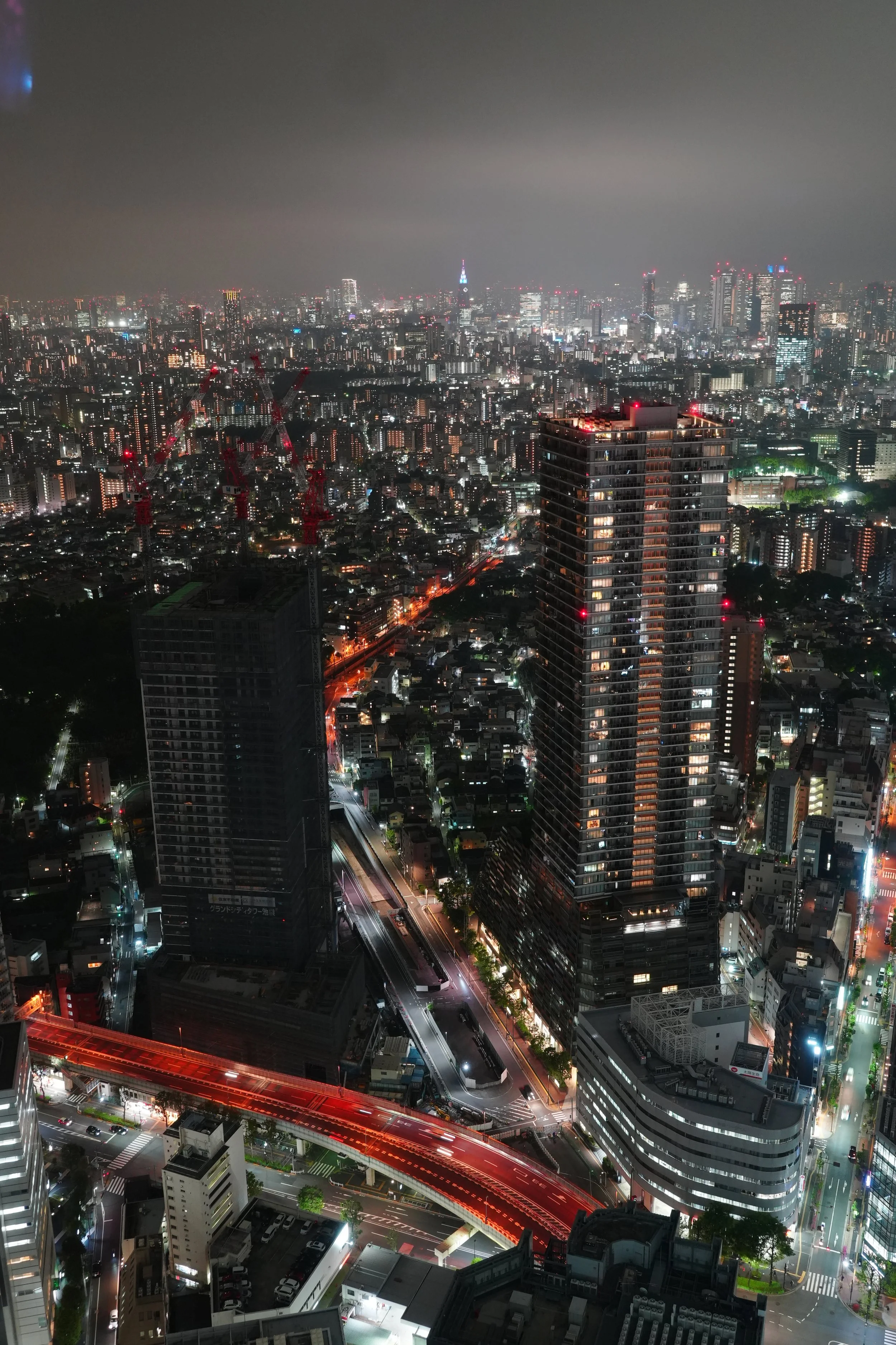 Night view of a cityscape with numerous illuminated buildings, skyscrapers, and construction cranes. The city is bustling with traffic, highlighted by red and white lights, and the sky is overcast.