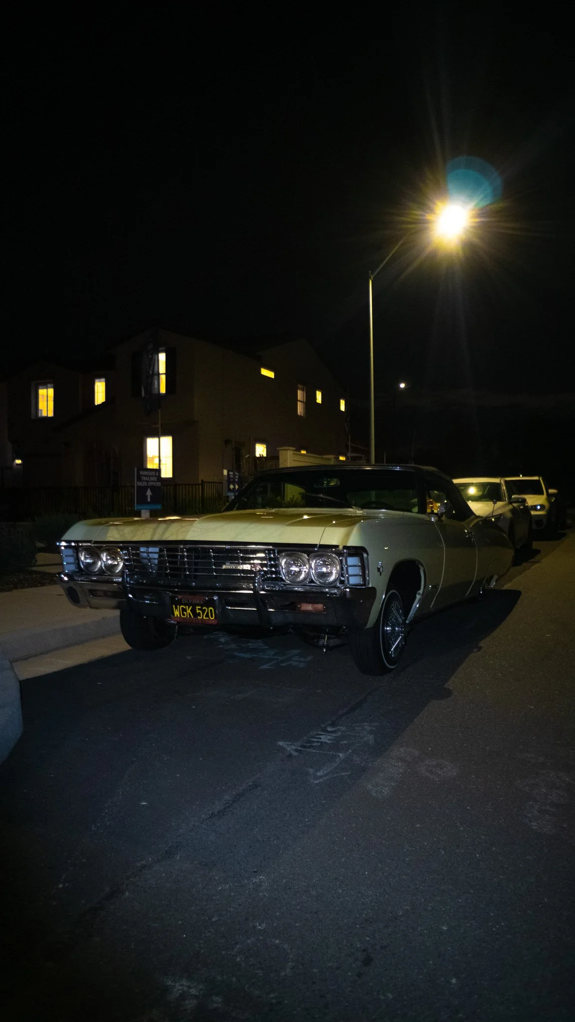 A vintage white Cadillac parked in a lot at night, illuminated by a bright streetlamp, with residential buildings in the background.