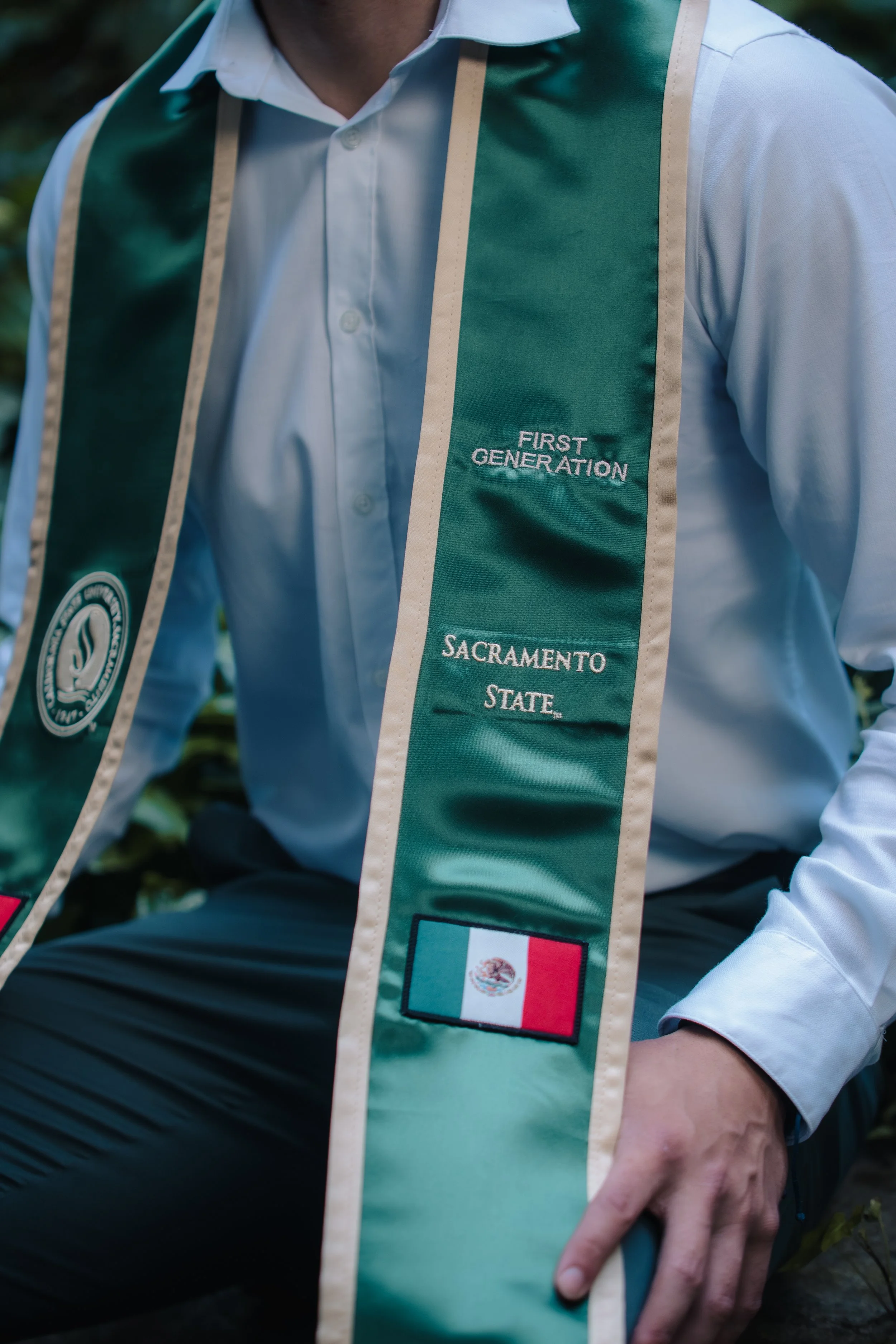 Close-up of a person wearing a white dress shirt with a green sash that reads "First Generation Sacramento State" and has a Mexican flag patch at the bottom. The person is sitting outdoors.