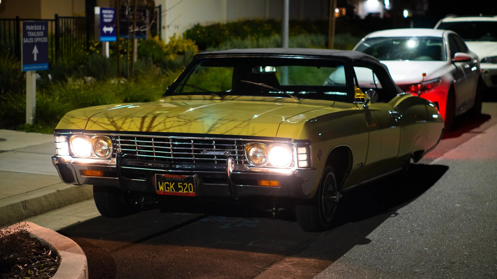 A yellow classic car with California license plate WGK 520 parked on a street at night, with its headlights on. There are other parked cars and signs in the background.