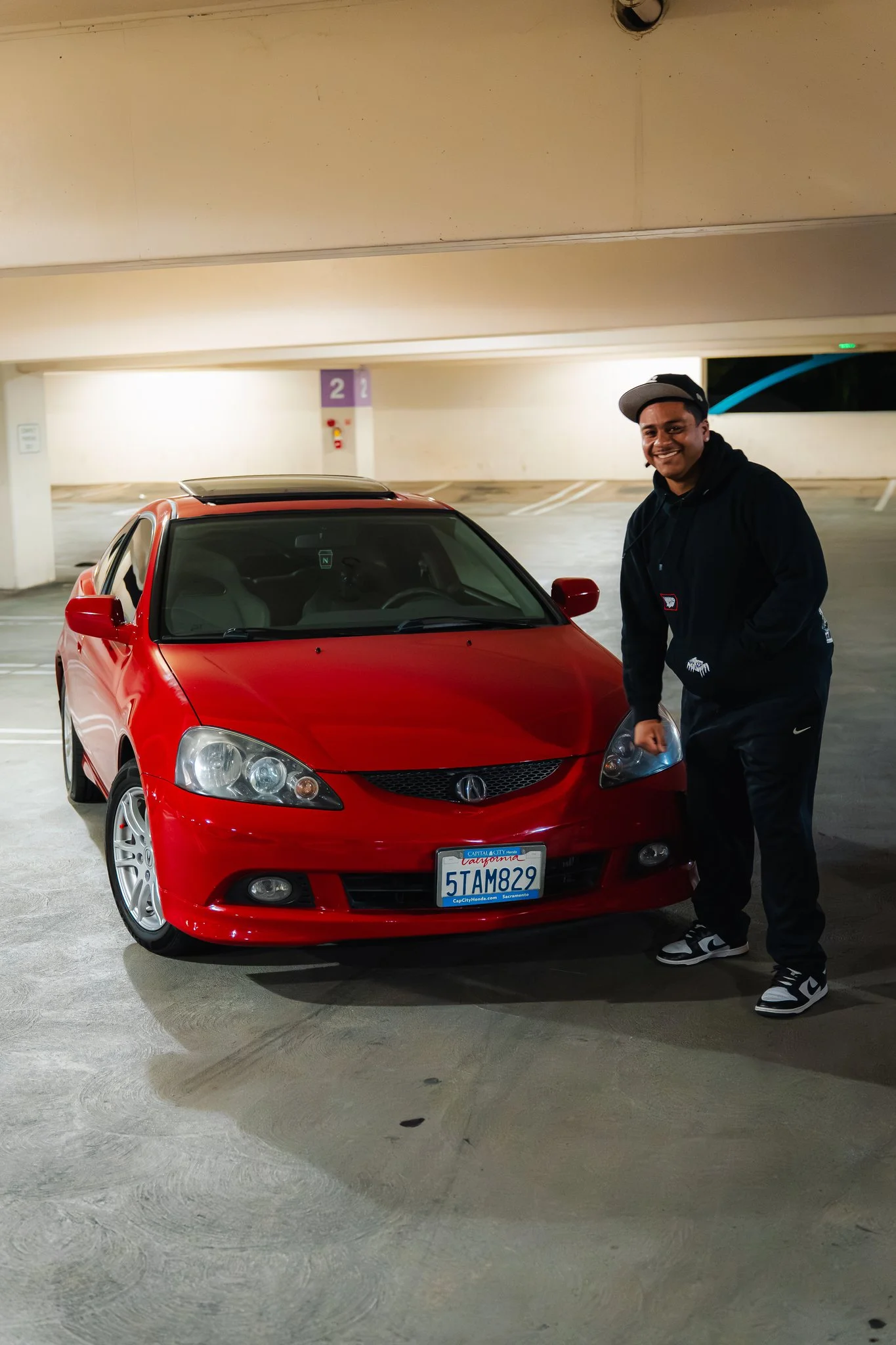 A young man smiling standing next to a red car with a California license plate in an indoor parking garage.