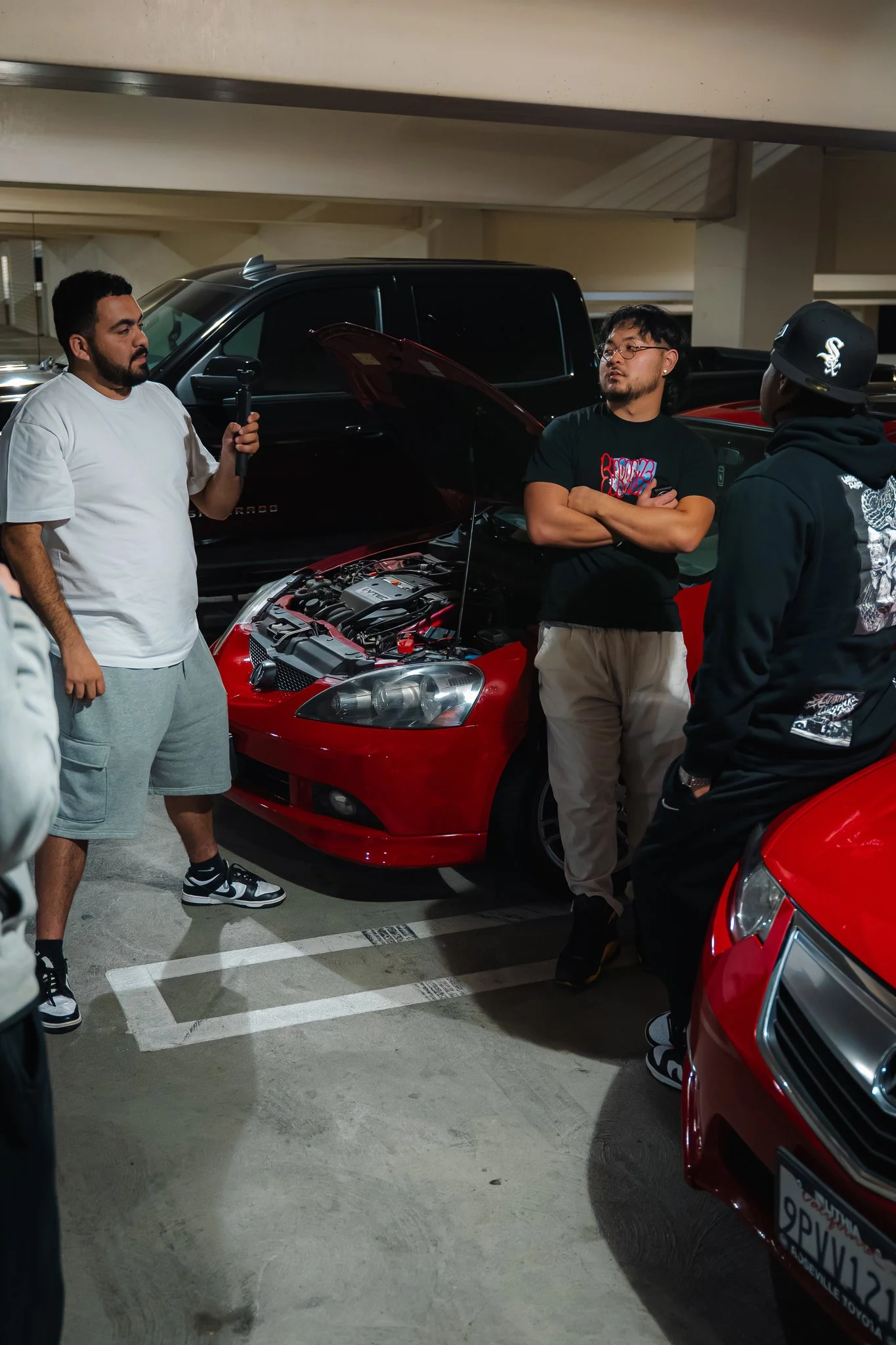 Three men are gathered around a red car with its hood open in a parking garage. One man, wearing a white t-shirt and shorts, is holding a microphone. Another man, with glasses and a black t-shirt, is standing with arms crossed, and the third man, wea