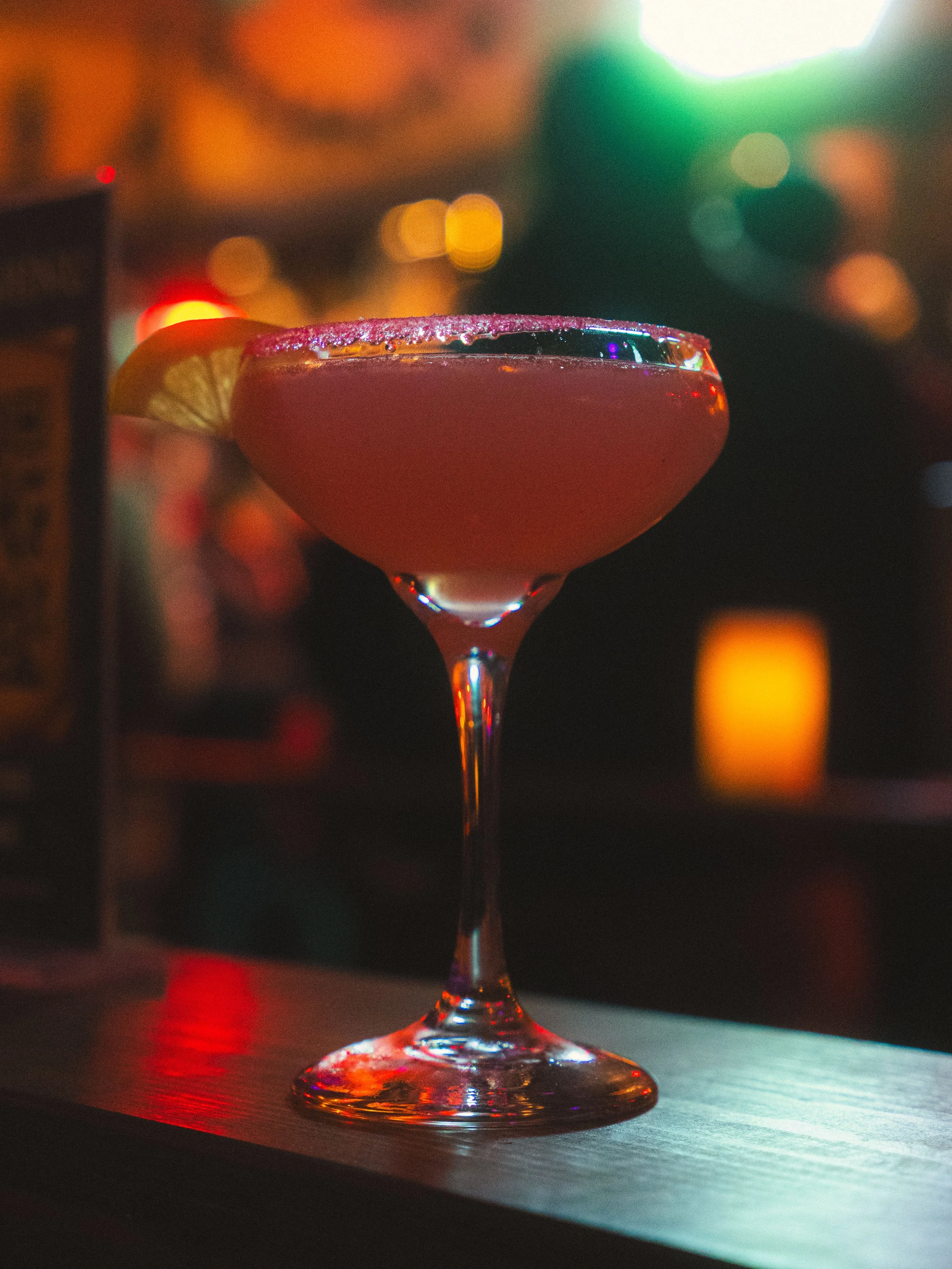 Close-up of a cocktail glass with a pink drink and a lemon wedge garnish, illuminated by colorful bokeh lights in the background.