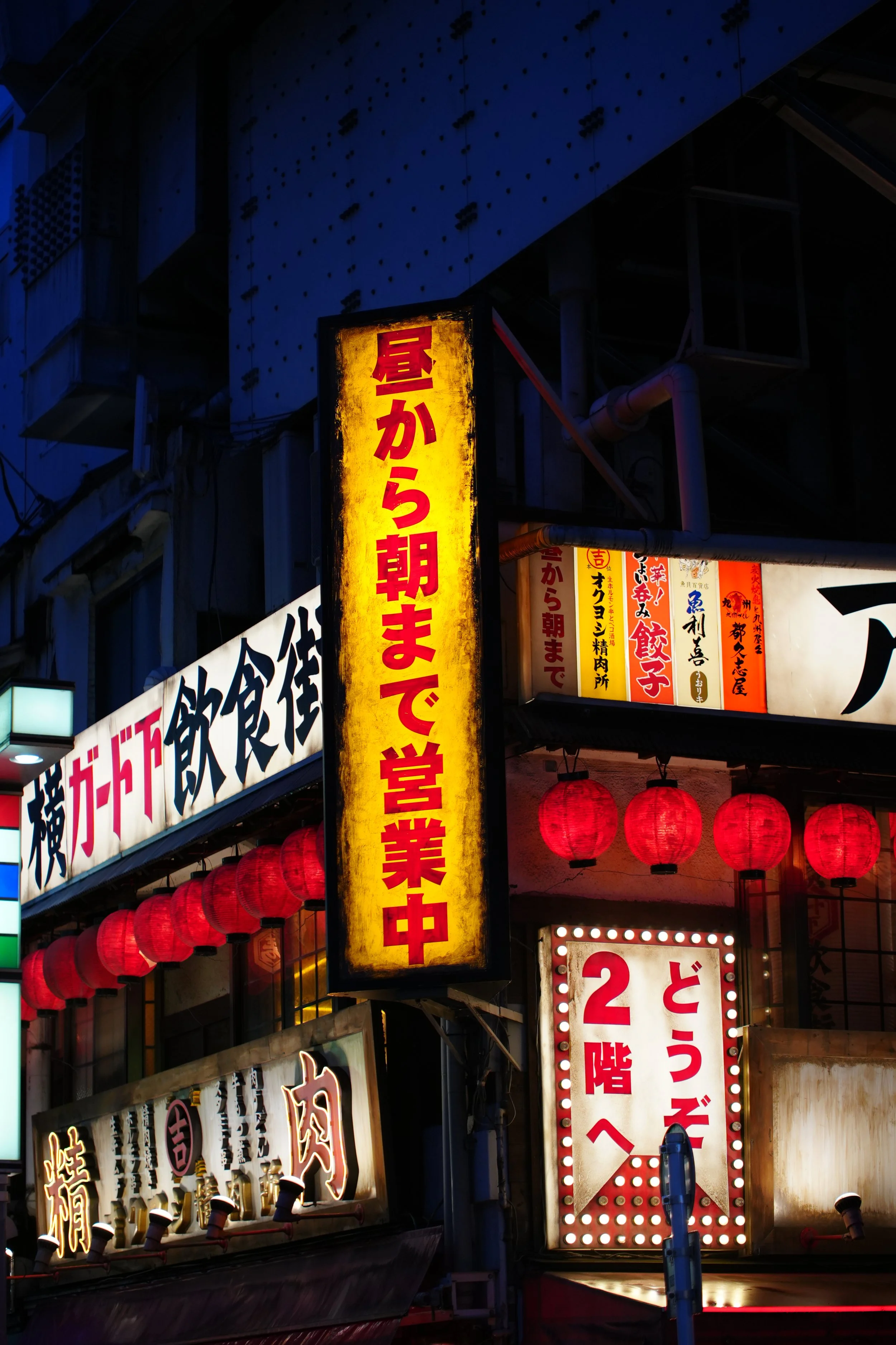 Night scene of a busy street with illuminated Japanese signs and red paper lanterns hanging outside buildings, indicating a lively area.