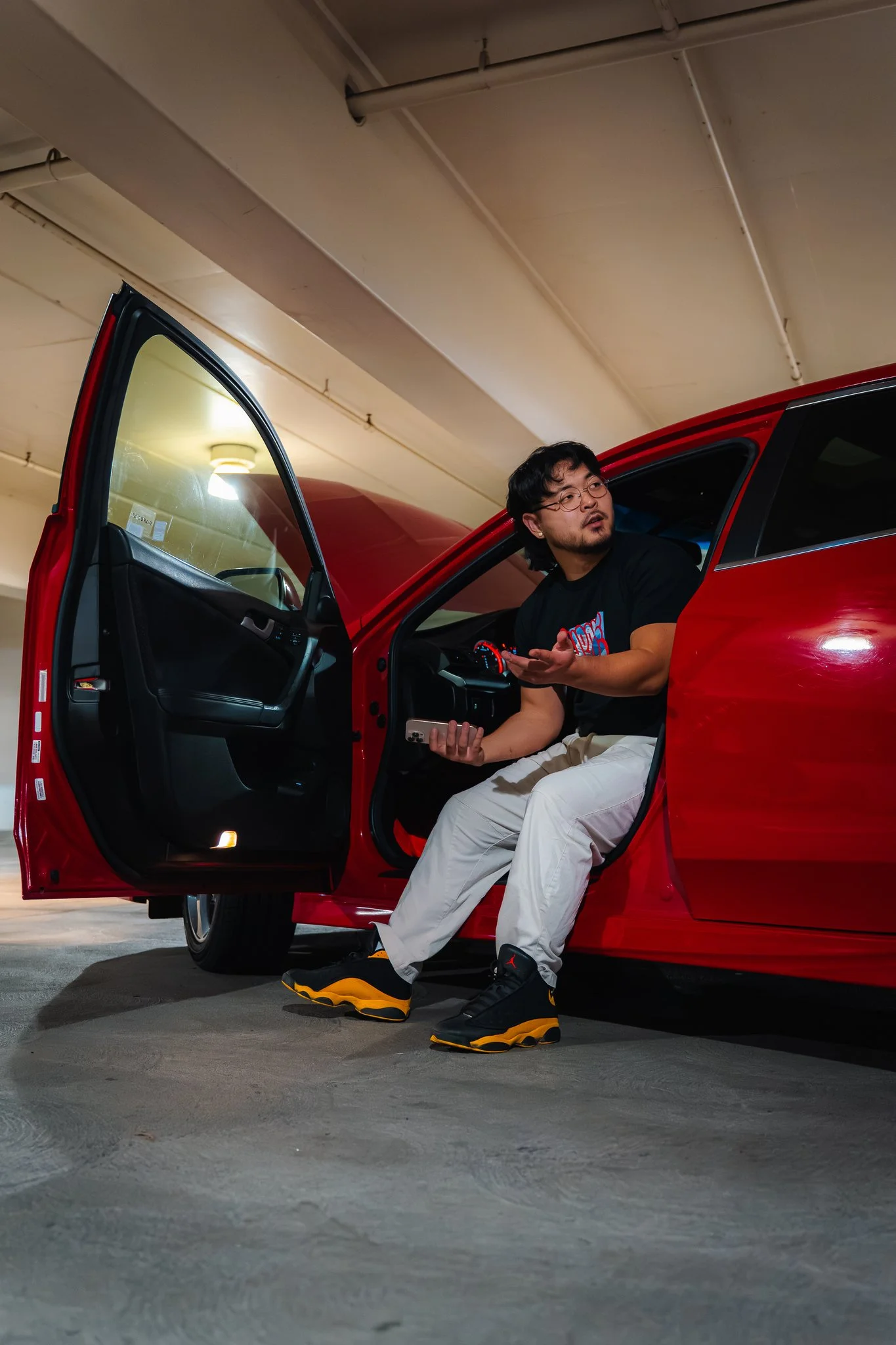 A man is sitting in the passenger seat of a red car with the door open, holding a smartphone, in an indoor parking garage with a beige ceiling and a bright ceiling light.
