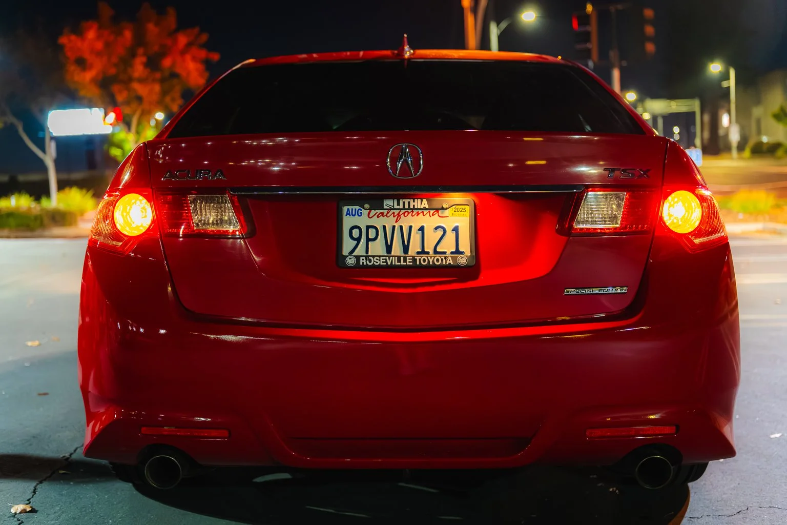 Rear view of a red Acura TSX car at night with illuminated taillights, showing a California license plate reading 9PVV121, parked on a street with lights and trees in the background.