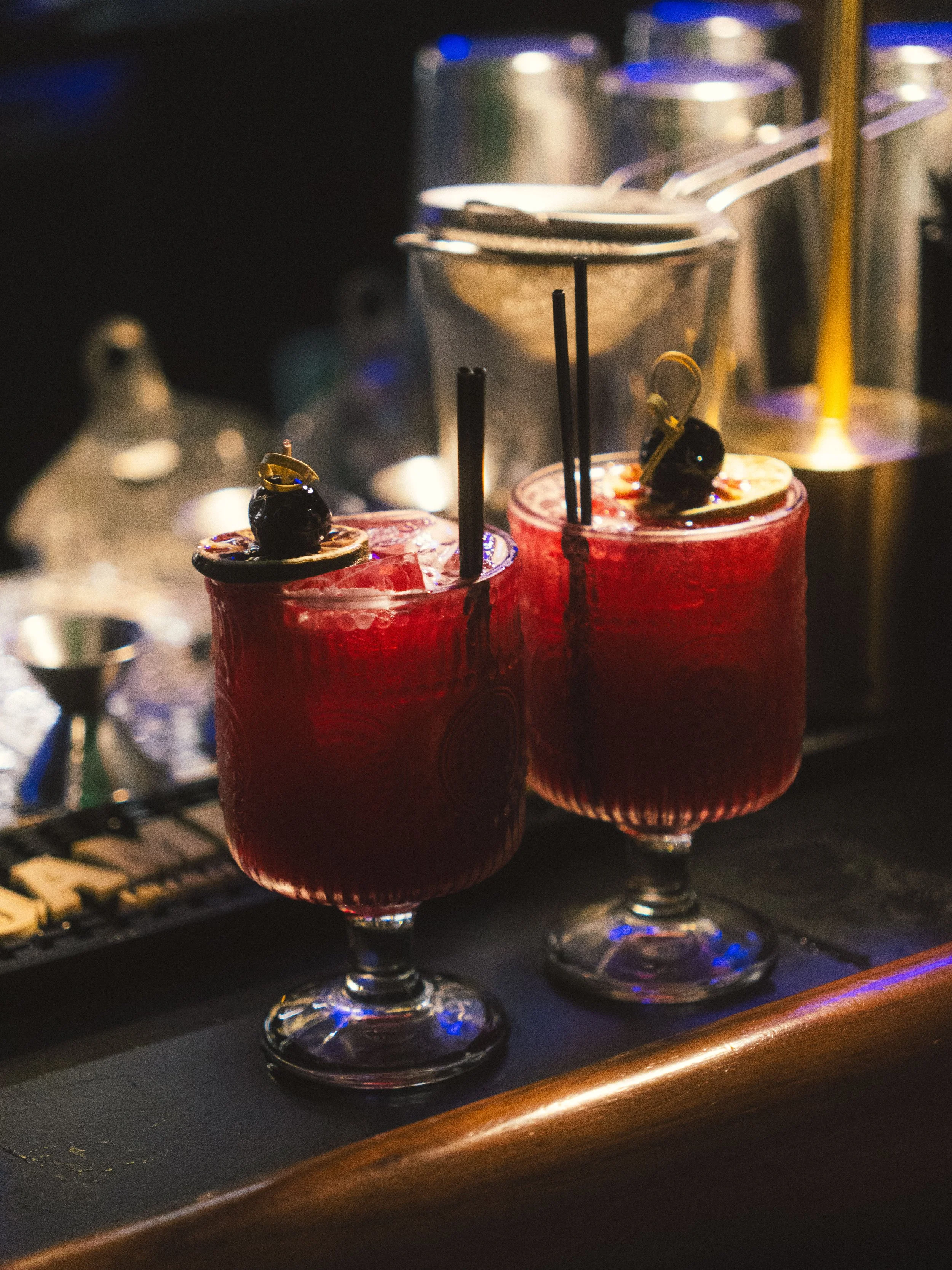Three red cocktails with black cherries and black straws on a bar counter in front of metal shakers.