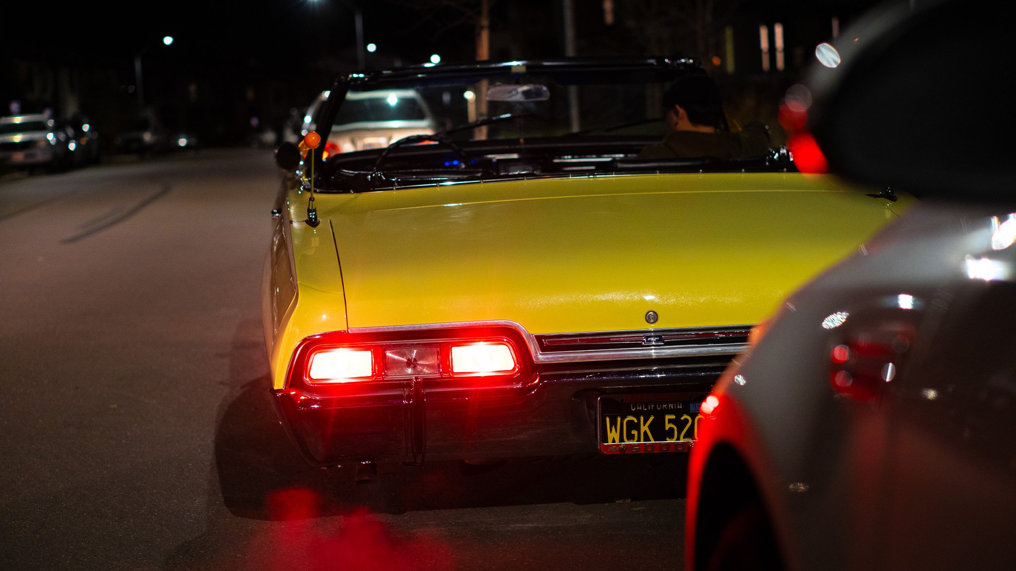 A yellow classic car with California license plate 'WGK 520' is driving on a street at night. The car has red tail lights and is surrounded by other vehicles, with some streetlights visible in the background.