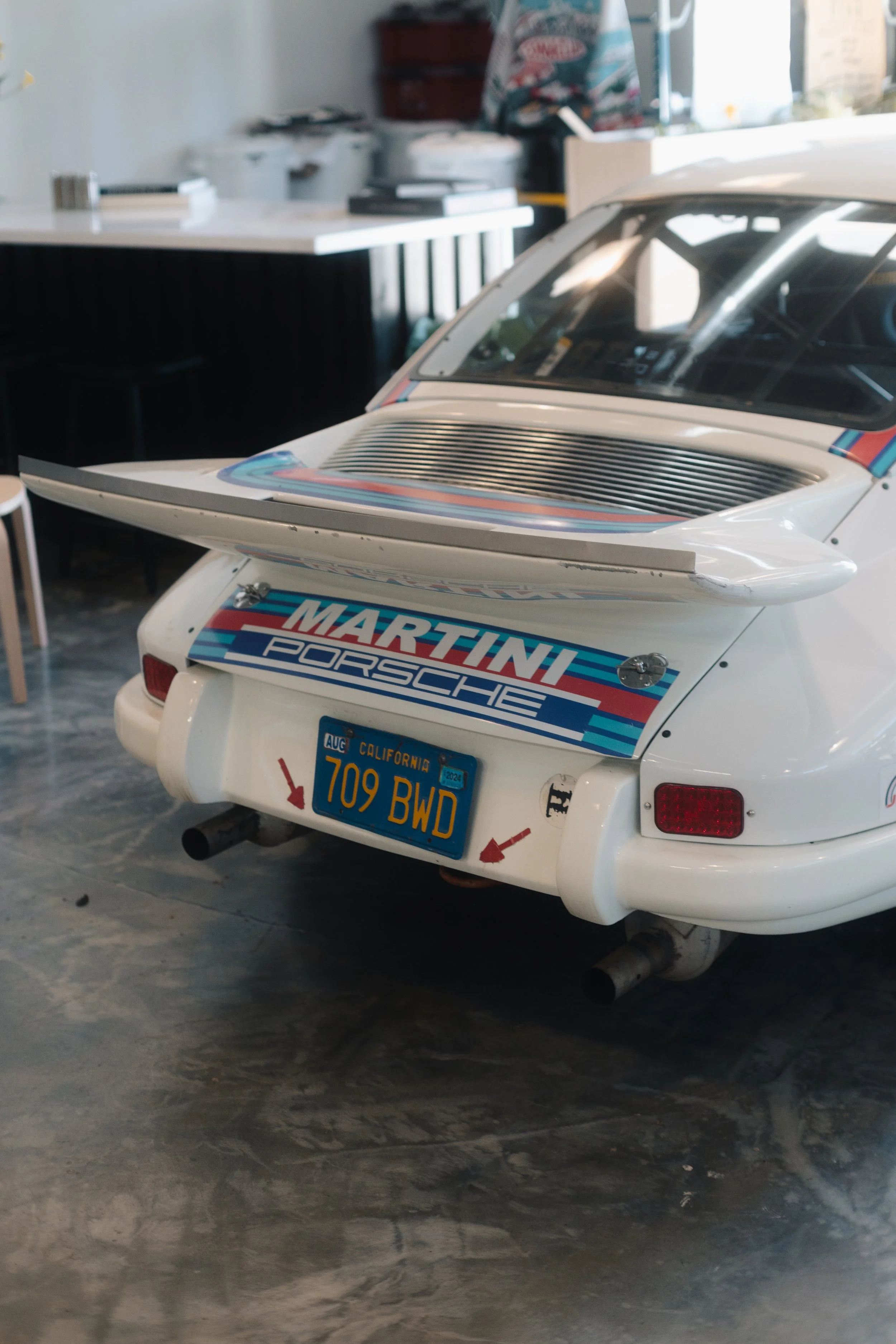 Rear view of a white Porsche race car with Martini racing livery, parked indoors on a concrete floor, with a blurred background of a kitchen or workshop area.