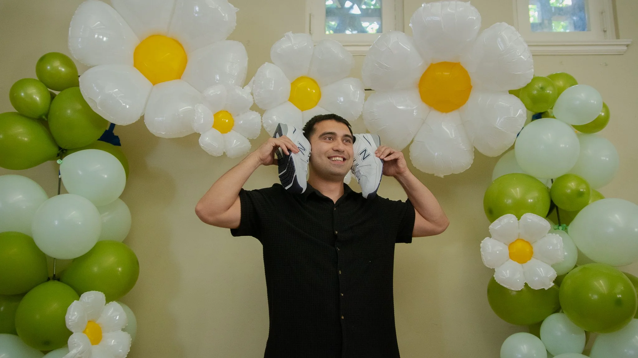 A man holding shoes on his ears stands in front of a backdrop of green and white balloons and large white and yellow flower-shaped balloons.