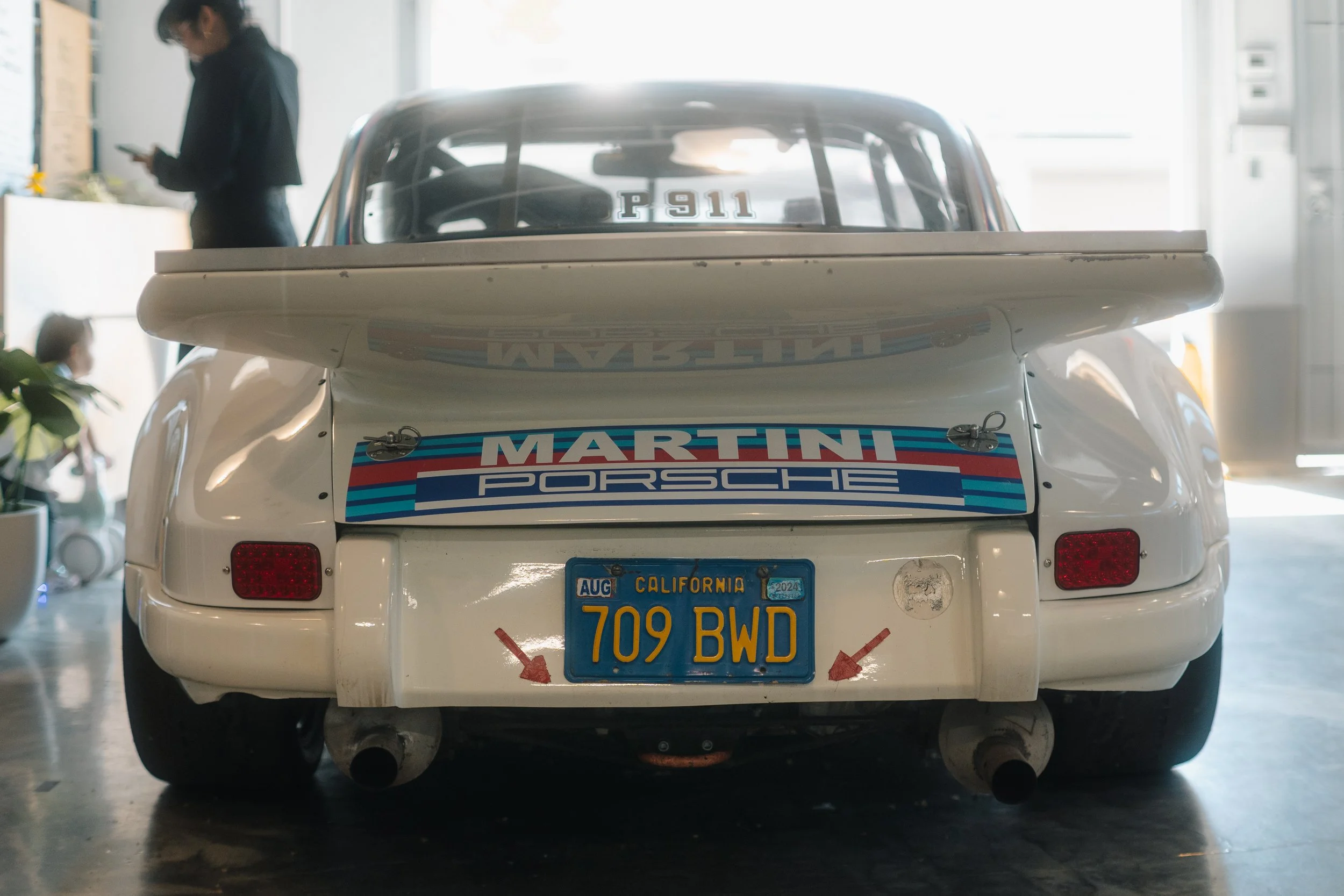 A rear view of a vintage white Porsche race car with racing decals, a license plate from California, and a large rear wing in a garage or showroom with two people in the background.