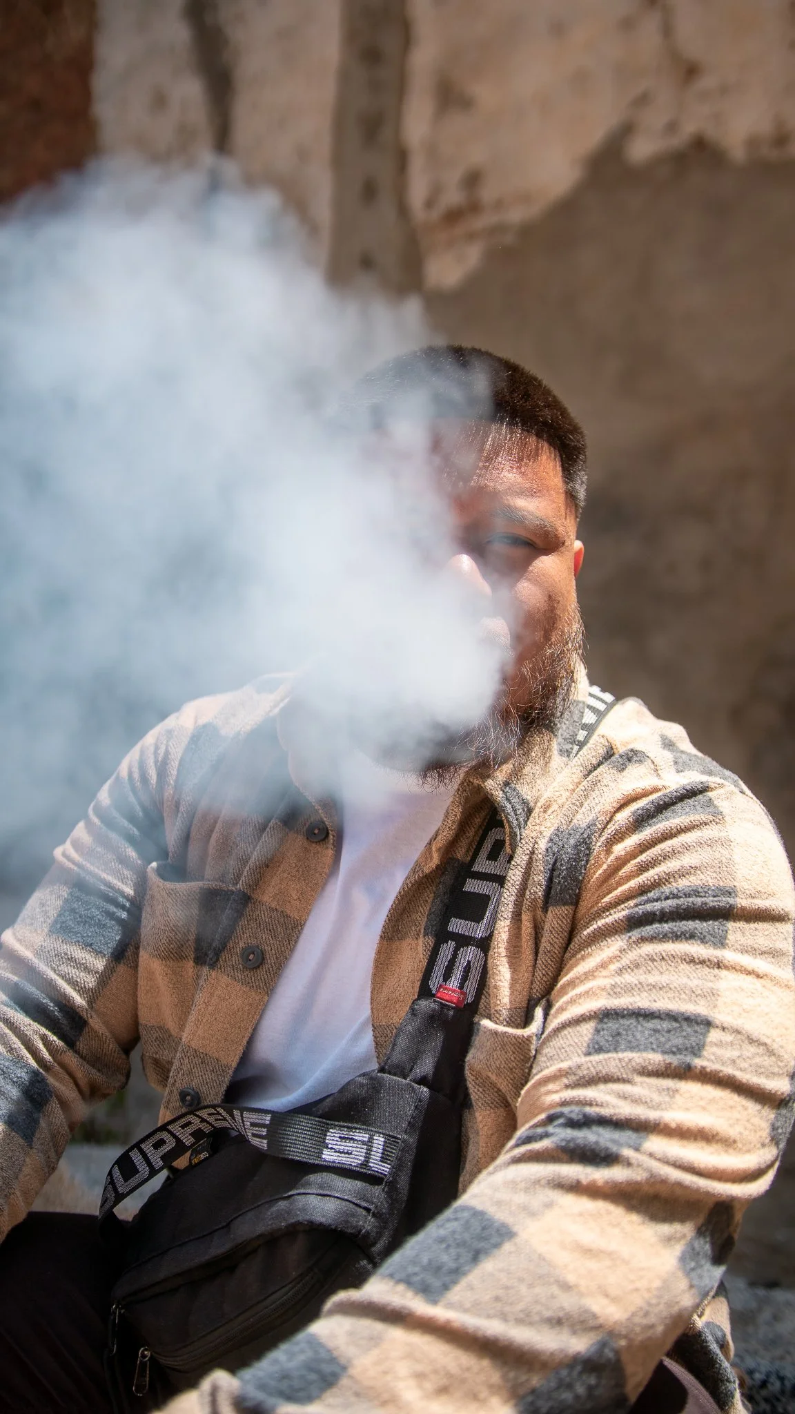 A man with a beard and short hair is sitting outdoors, partially obscured by a cloud of smoke or vapor.