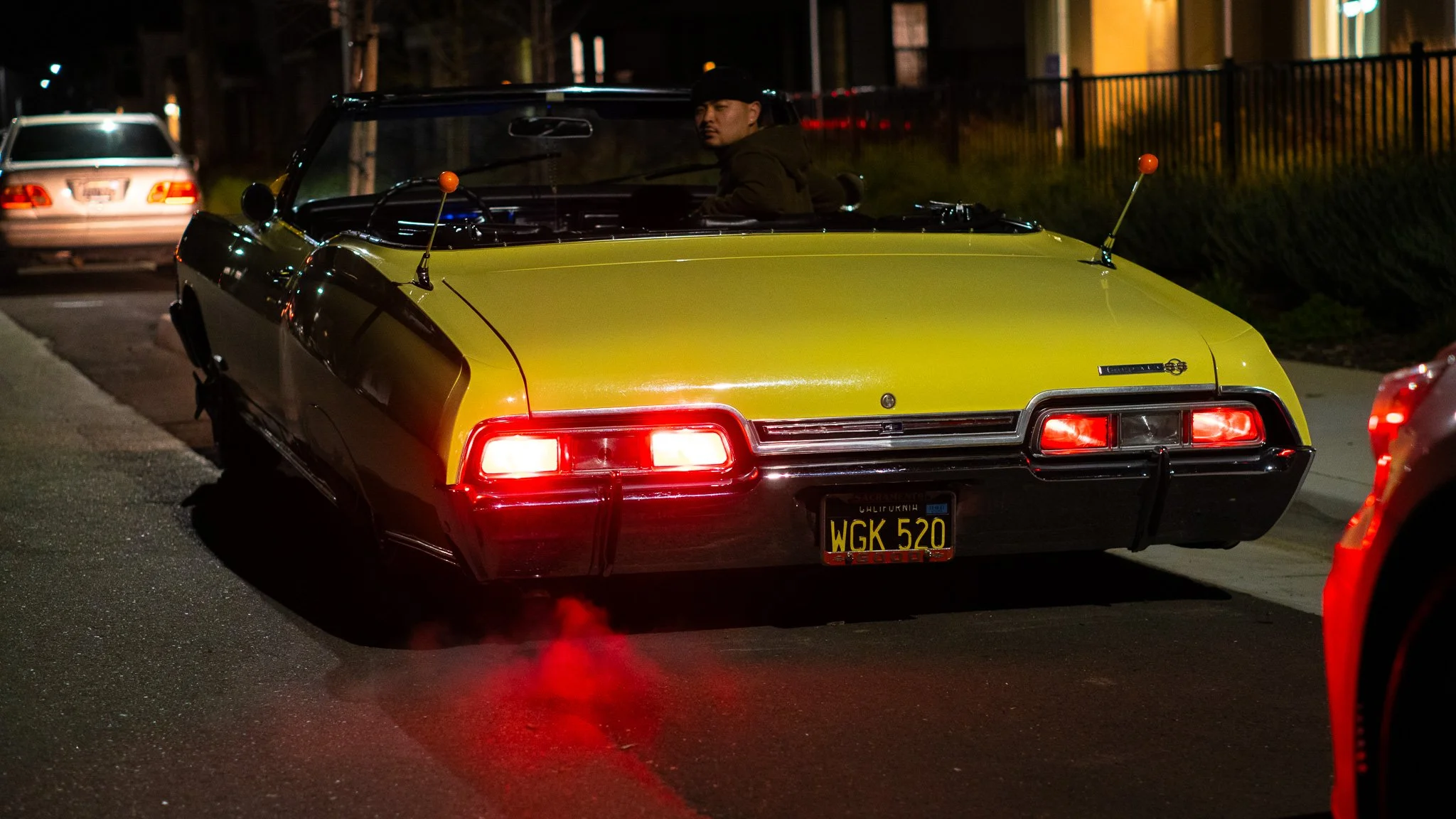 A yellow vintage car with a black roof is parked on a street at night, with a person sitting inside looking to the side. The car has orange antenna balls on the front and rear. There is another car parked behind it and a dark, fenced area in the back