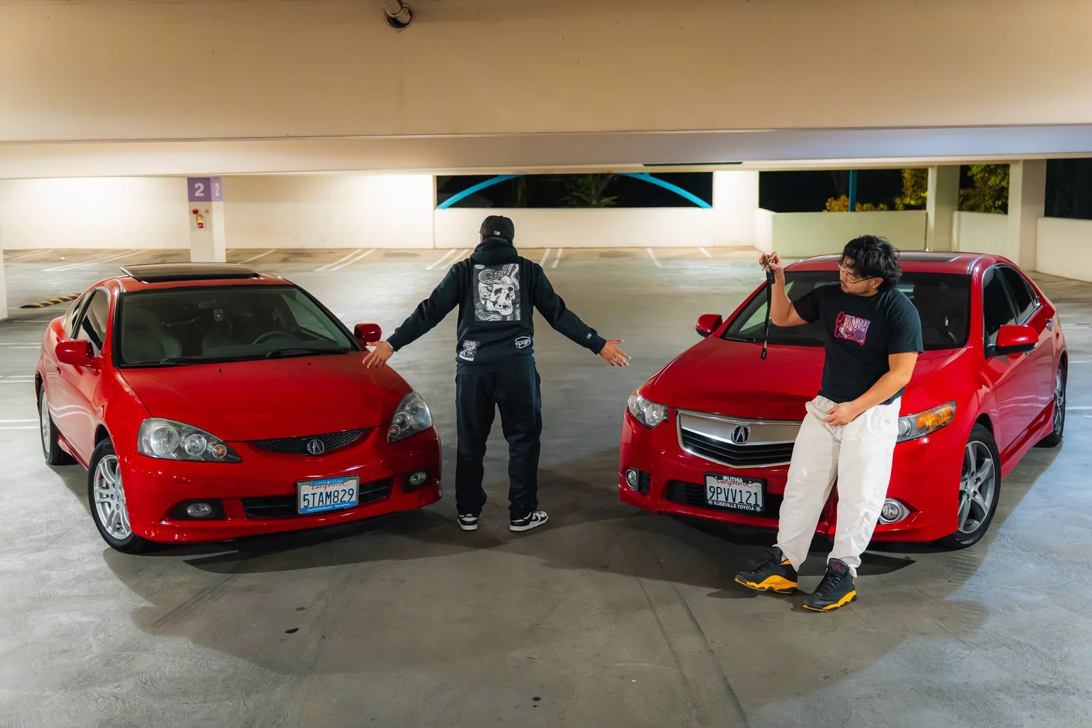 Two young men in a parking garage posing with two red cars facing each other. The man on the left has his arms outstretched, wearing a black hoodie with a graphic on the back, and the man on the right holds sunglasses in his hand, wearing a black t-s
