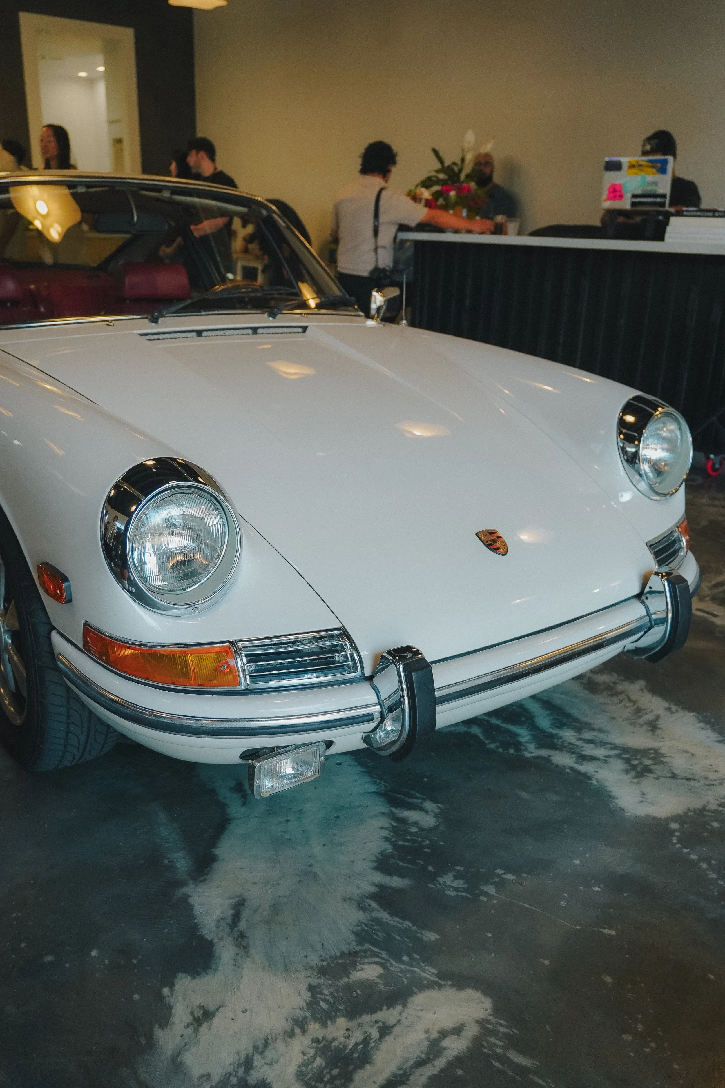 A white classic Porsche sports car parked indoors, with people in the background at a reception desk and floral arrangements.