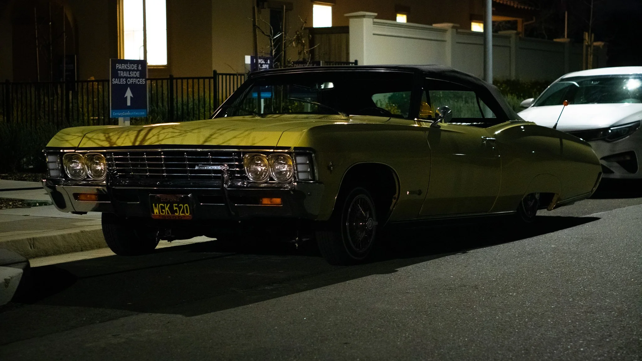 A vintage green and black car parked on the street at night, with another modern white car parked behind it. There is a sign indicating directions to sales offices.