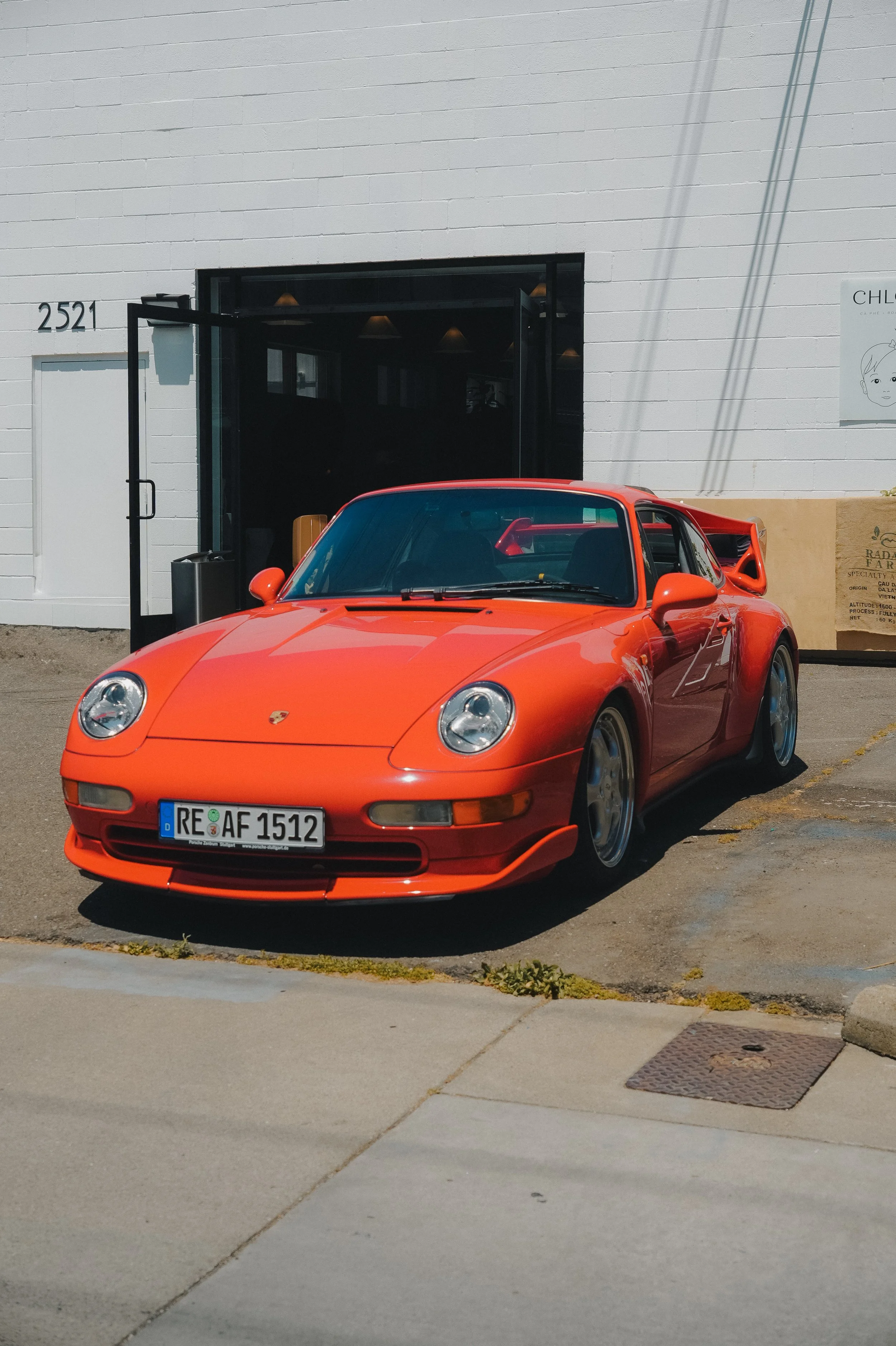 An orange sports car parked in front of a white building with the address 2521. The car has a sleek design, alloy wheels, and a rear wing spoiler.