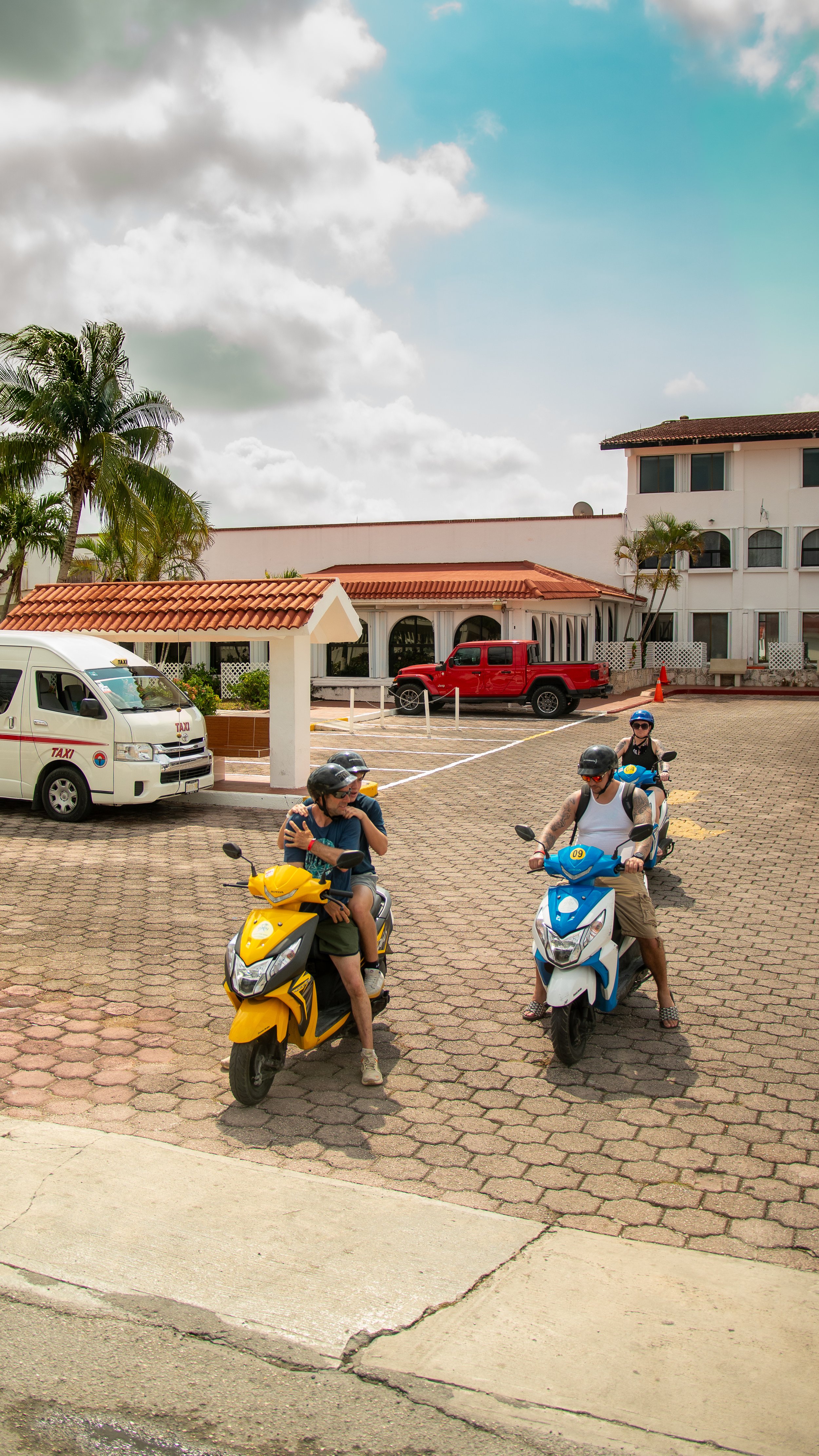 People riding scooters in a parking lot with a white building, palm trees, and a partly cloudy sky in the background.
