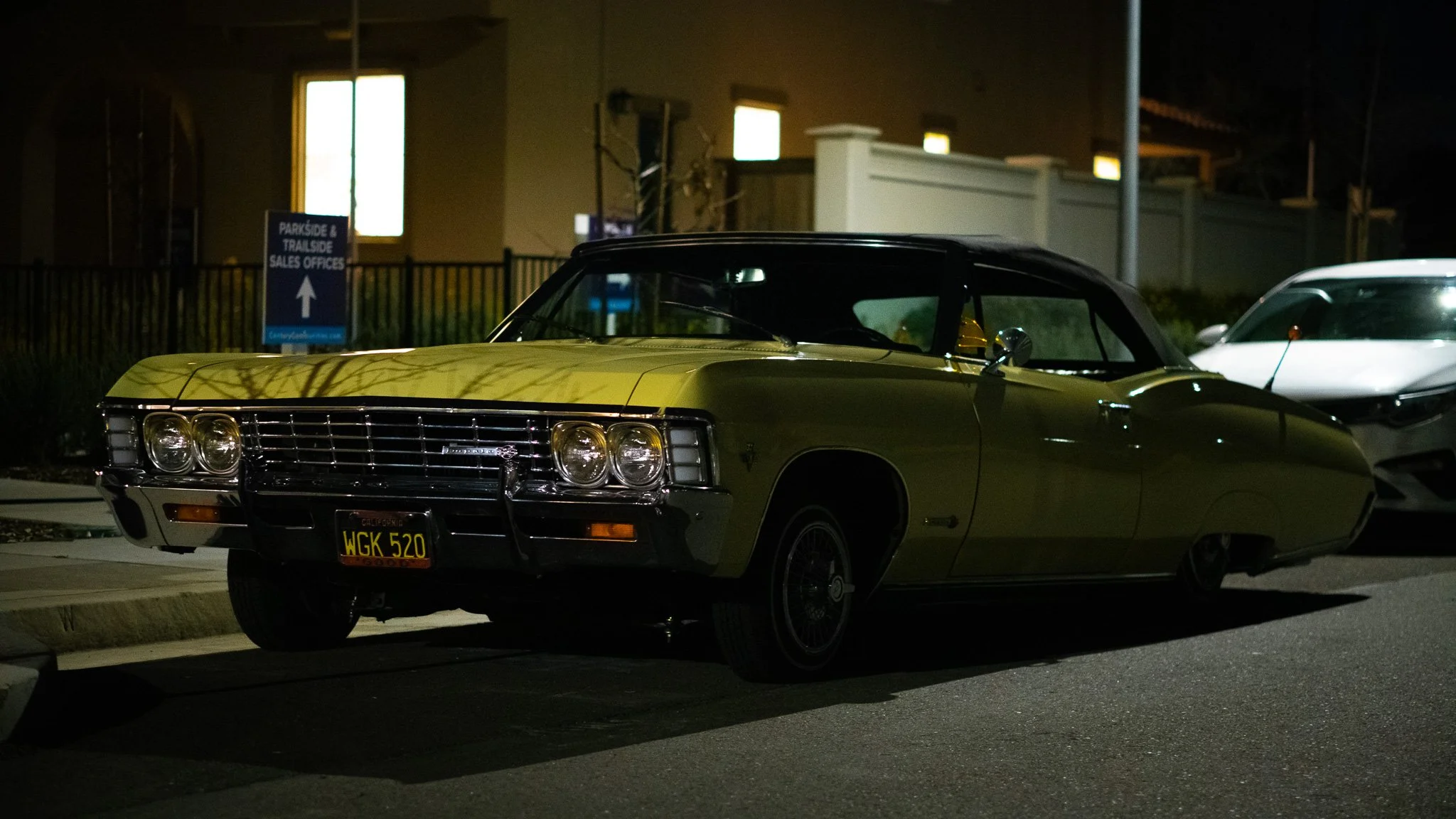 A vintage yellow convertible car parked on a city street at night, with another modern white car beside it, and a sign for Paradise & Trasco sales offices in the background.