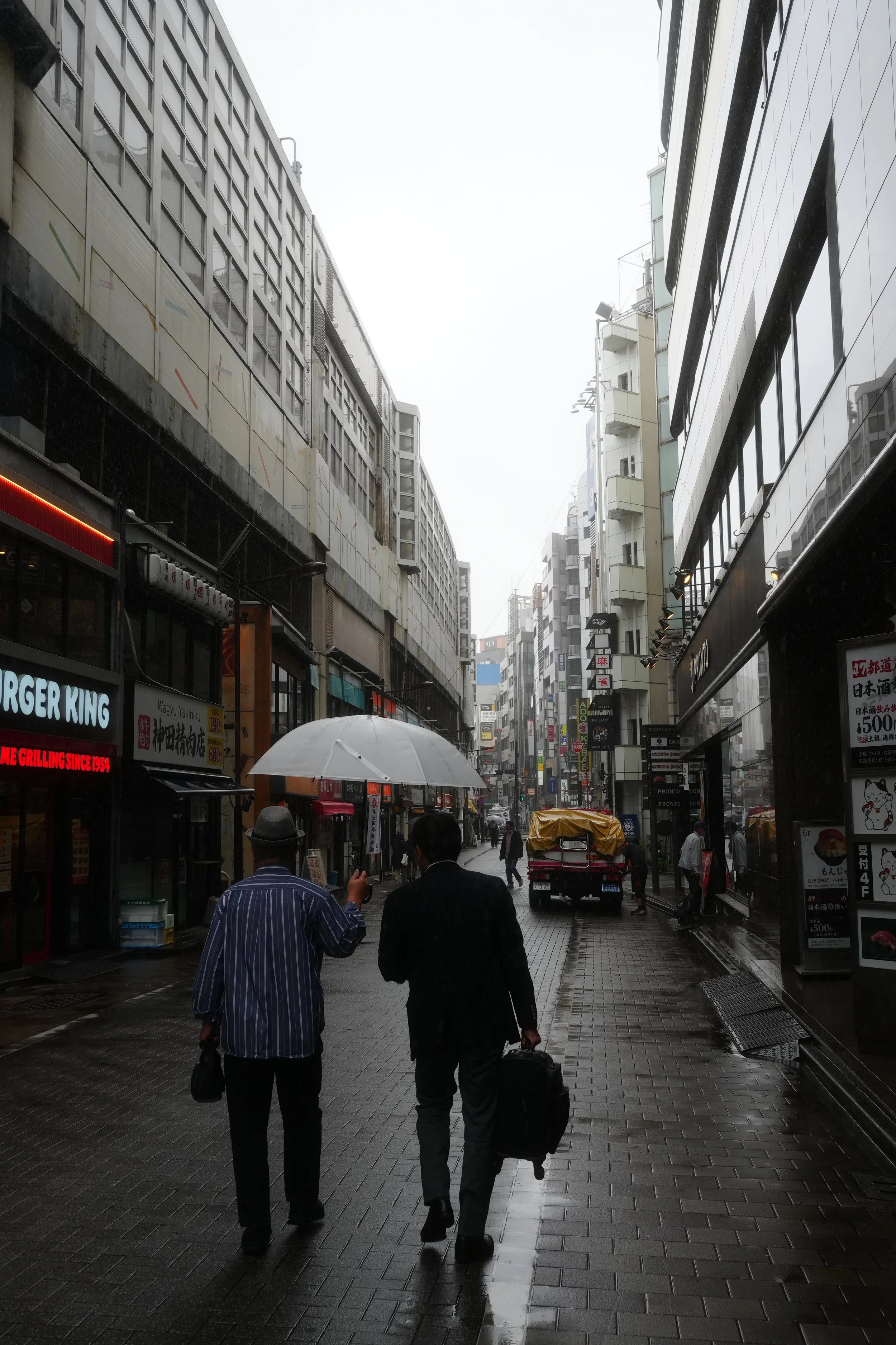 Street scene in a city on a rainy day with people walking on the sidewalk, some holding umbrellas. Buildings line both sides of the street, and there are various signs and storefronts visible.