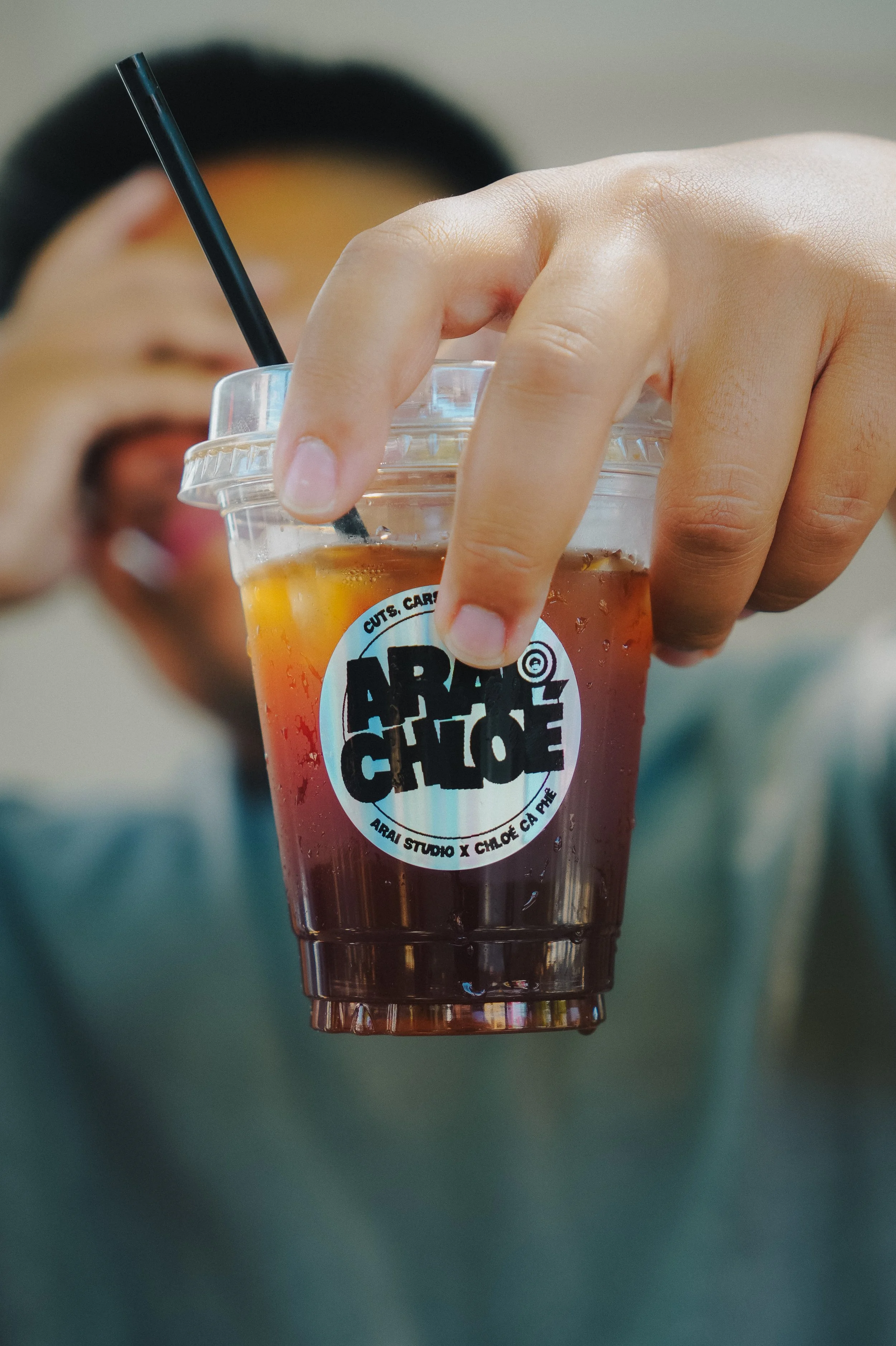 Close-up of a hand holding a cup of iced coffee with a straw, featuring a sticker that reads 'ARAI STUDIO x CHLOE CHLOE' with blurred person in background.