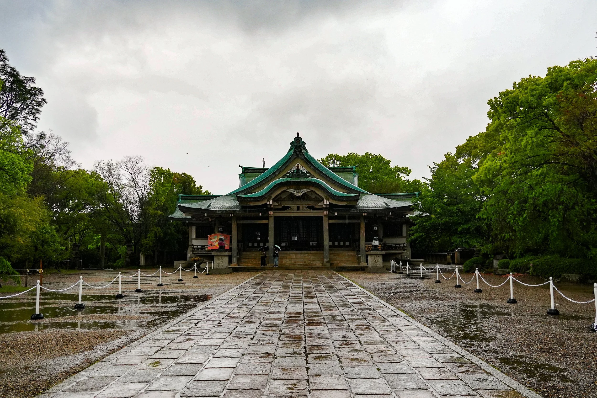 A traditional Japanese shrine building with a green roof surrounded by lush green trees on a cloudy day. The pathway leading to the shrine is made of stone tiles, and there are people standing at the entrance under umbrellas.