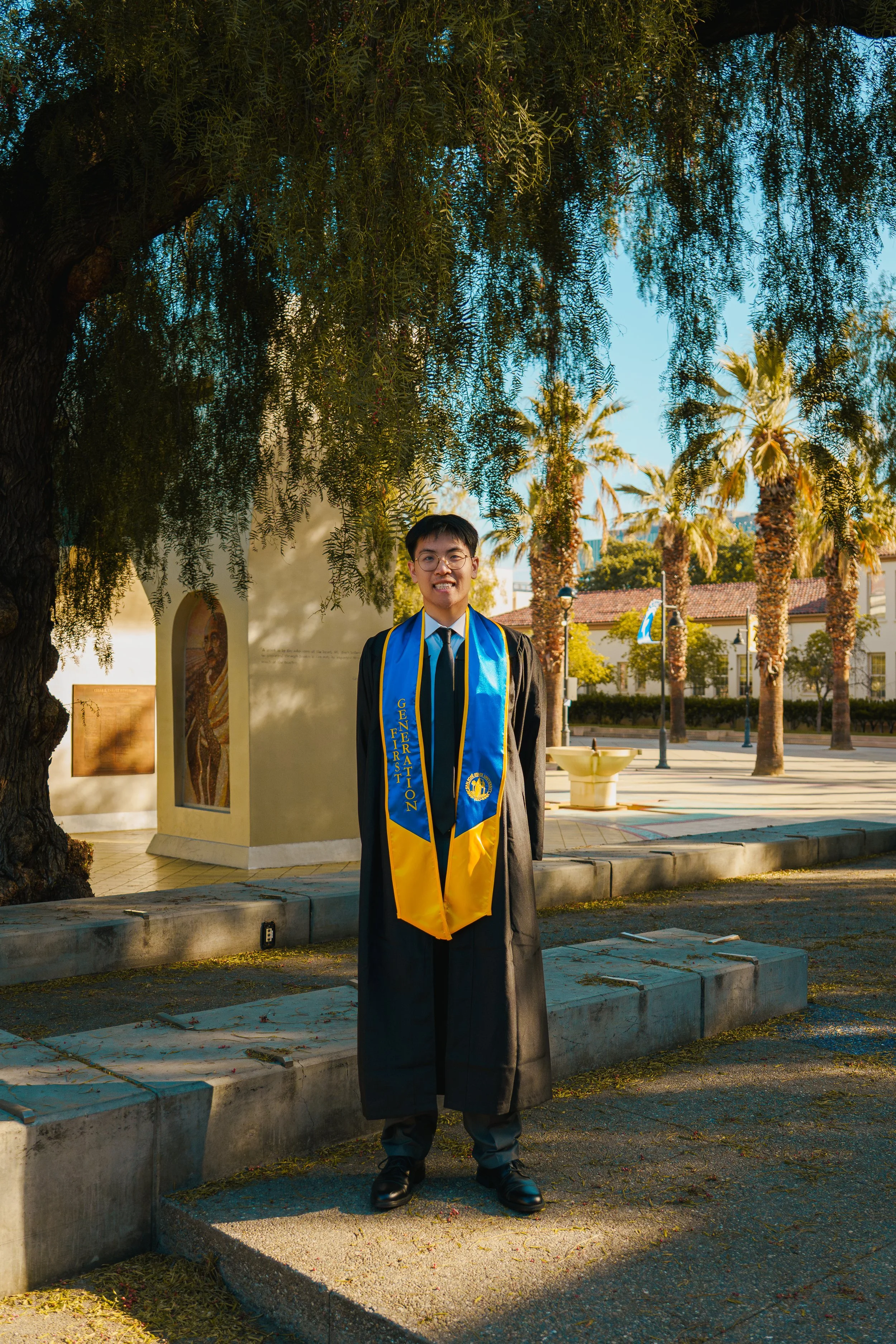 A young man in graduation attire standing outdoors under a large tree with palm trees in the background. He is smiling and wearing a blue and yellow sash with the words 'First Generation' on it.