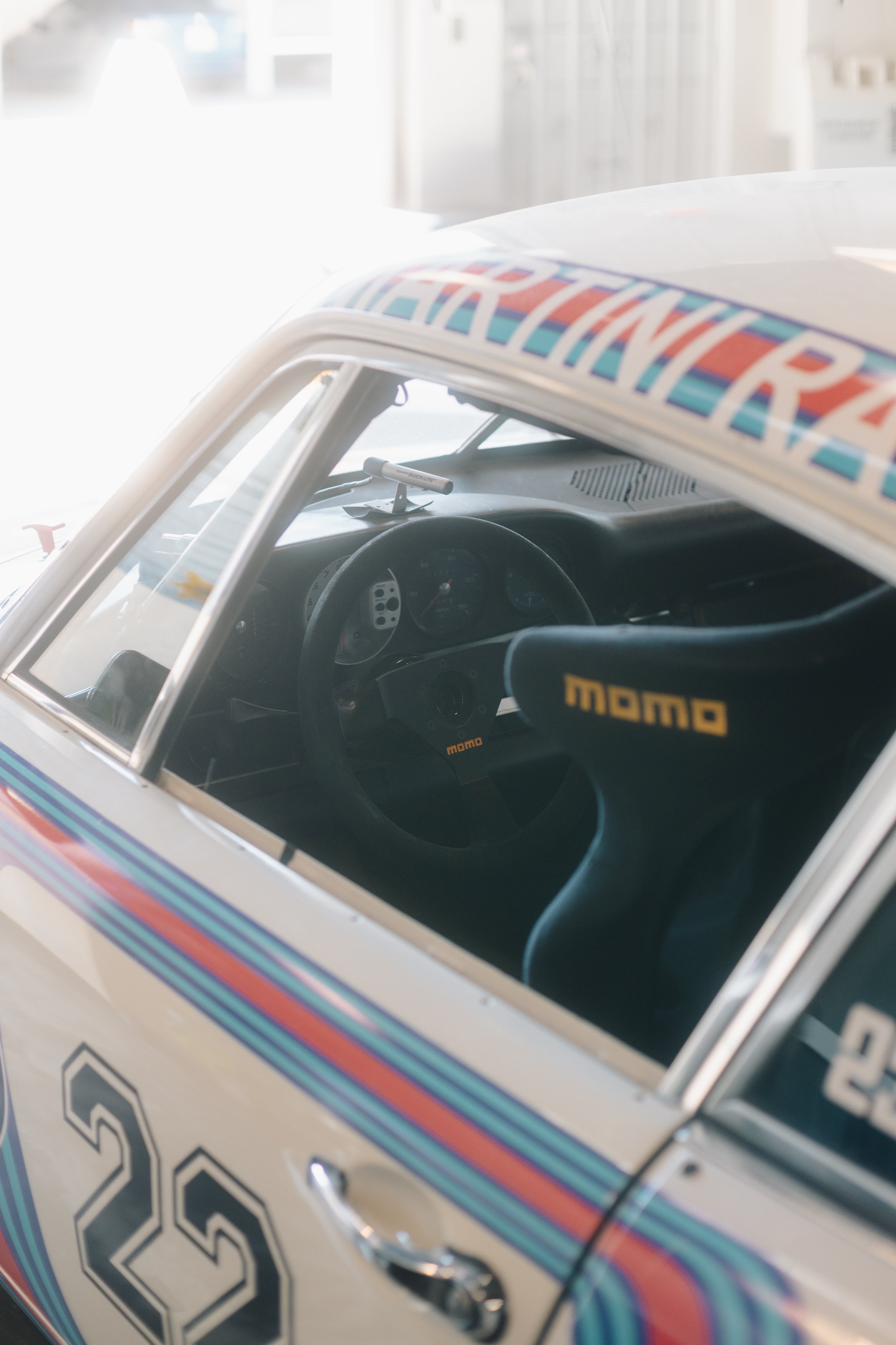 Close-up of a race car displaying the number 28, with visible interior including steering wheel labeled MOMO and dashboard, through a partially transparent windshield. The car has a colorful stripe design on the exterior.