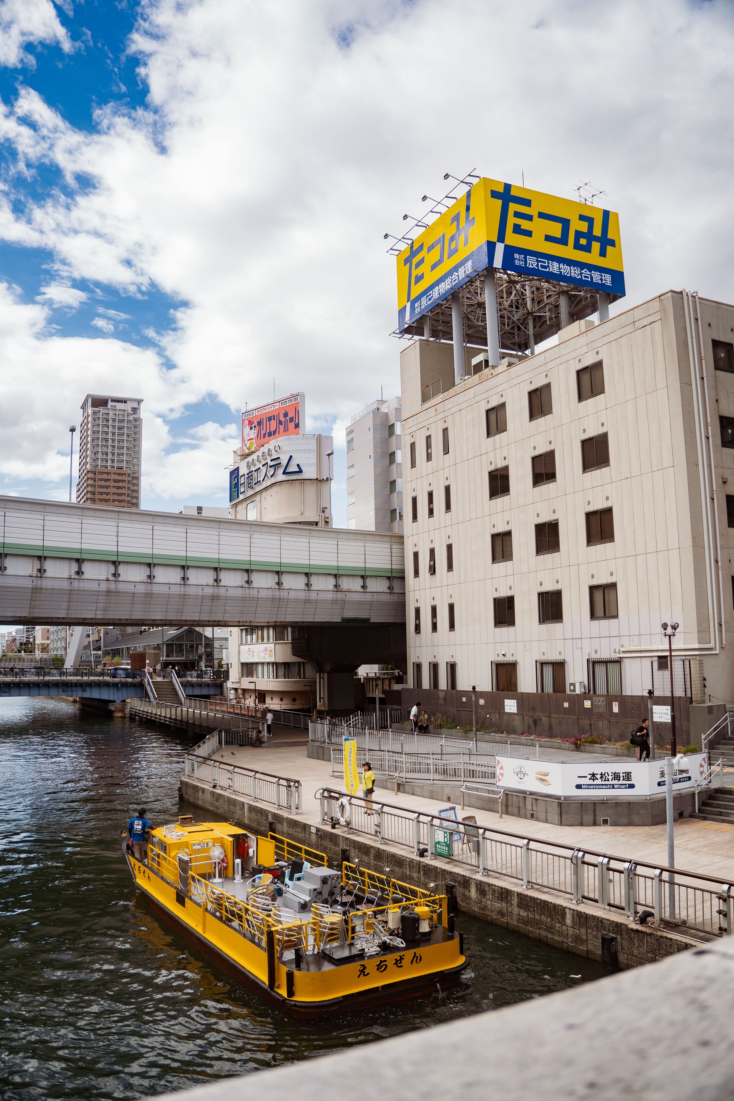 A yellow sightseeing boat named 'えちぜん' on a river in a city, with people on a waterfront promenade, tall buildings, and billboards in the background under a partly cloudy sky.
